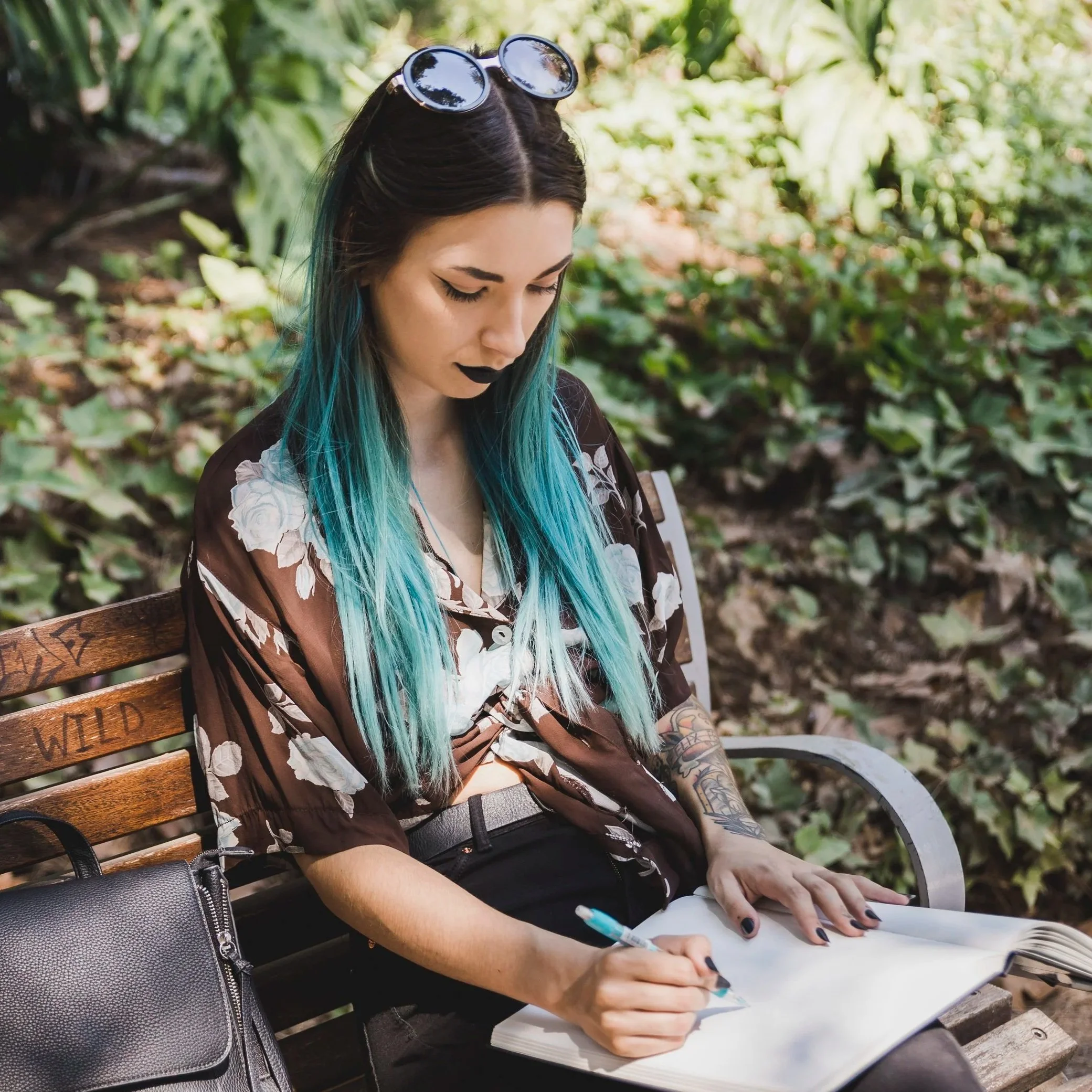 A young woman with long turquoise hair, wearing sunglasses on her head, black lipstick, and a floral blouse, sitting on a wooden park bench outdoors, writing in a notebook with a pen.