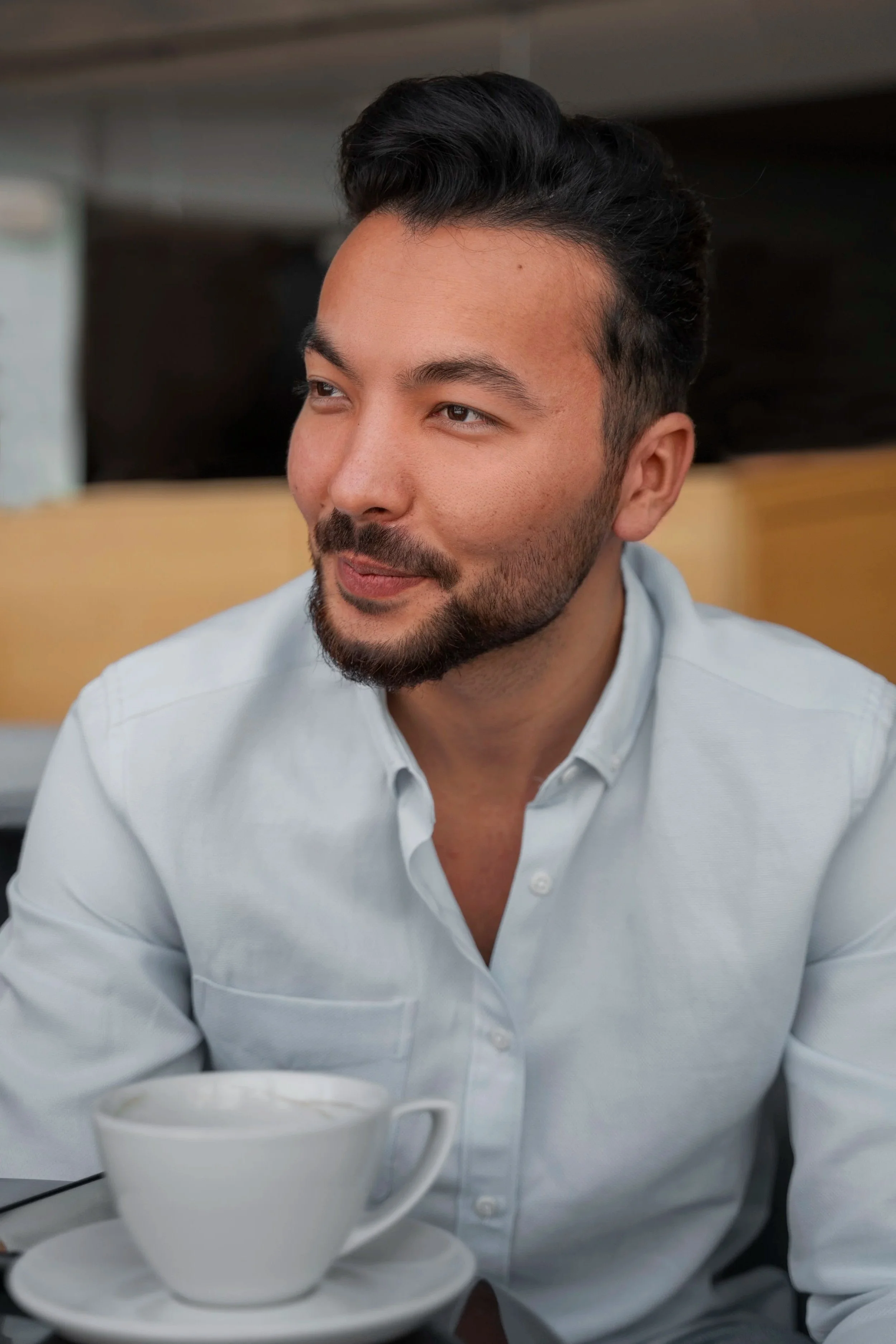 A man with dark hair and a beard sitting at a table with a cup of coffee, wearing a light-colored button-up shirt, in a cozy indoor setting.