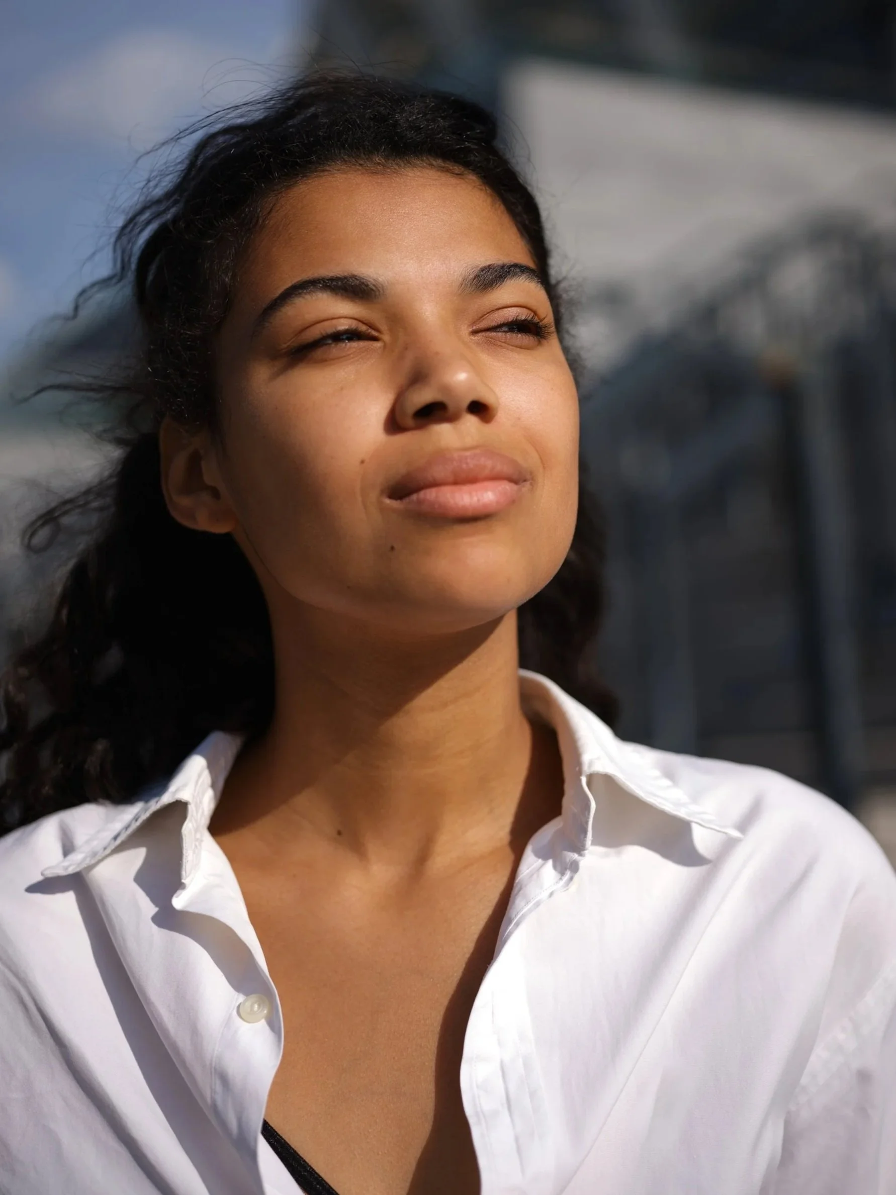 A woman with dark hair and a light complexion looking towards the sunlight, wearing a white shirt, with a blurred background.