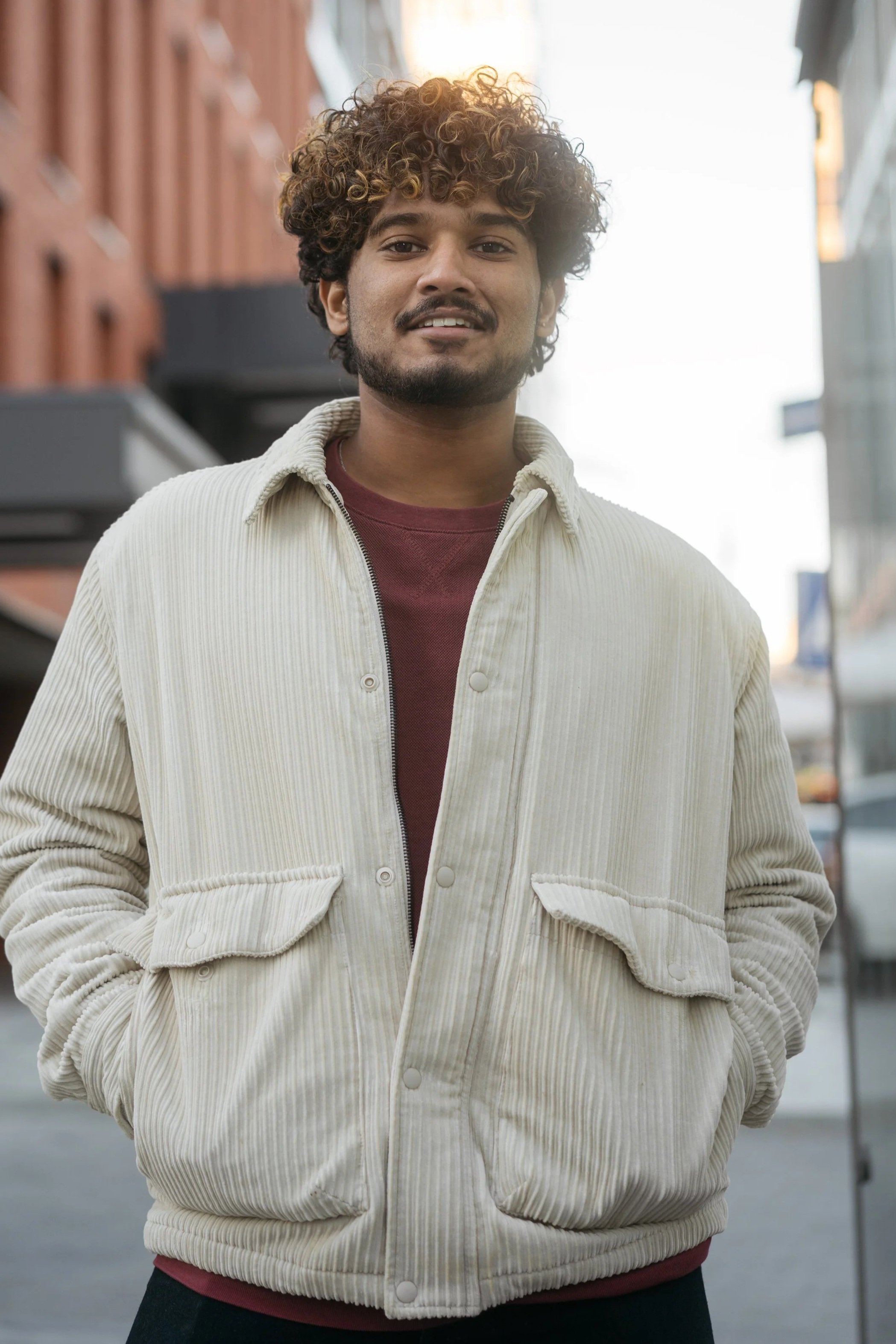Man with curly hair smiling outdoors in an urban setting, wearing a cream-colored corduroy jacket over a red shirt.