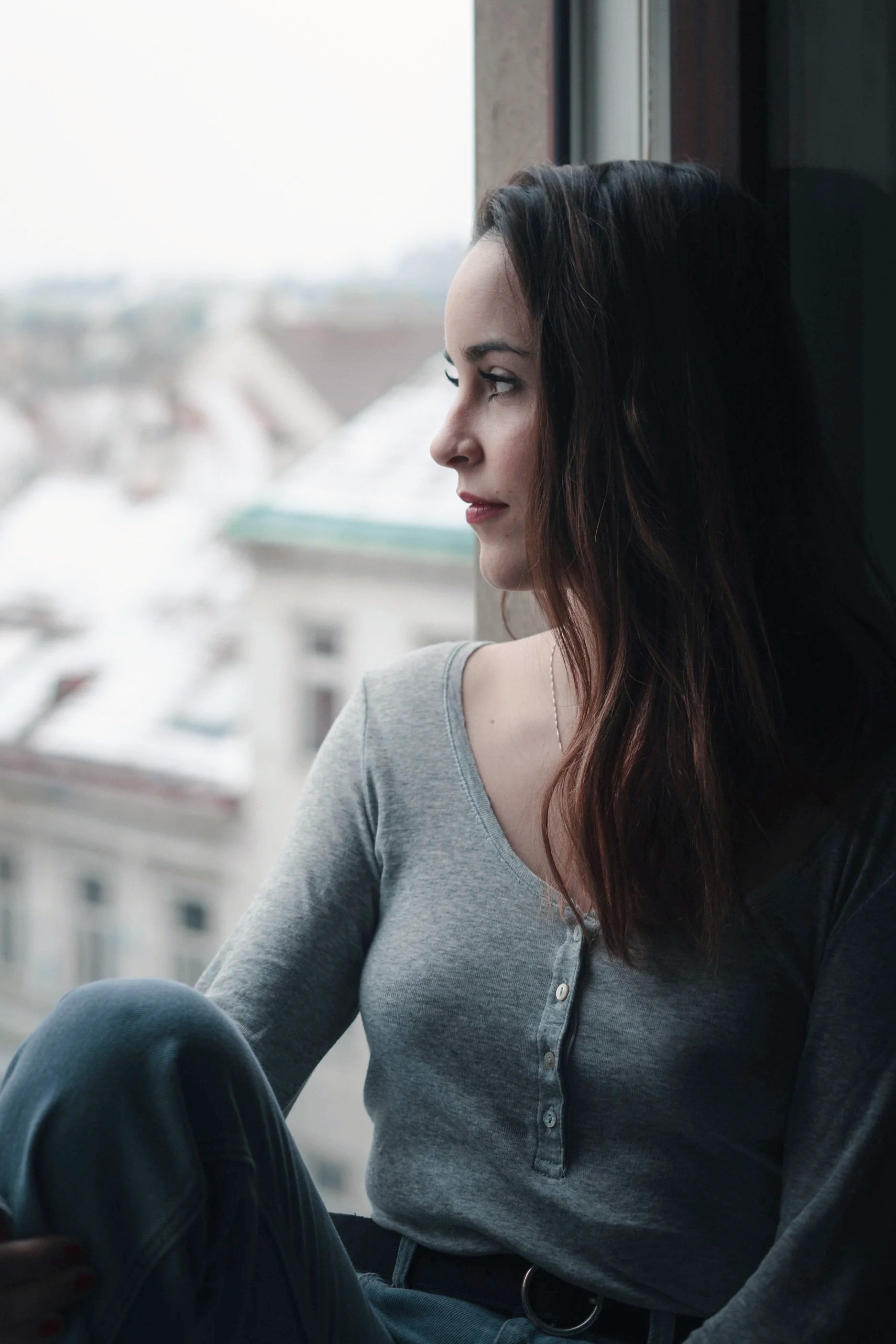 A woman with dark hair sitting by a window, looking outside on a snowy day.