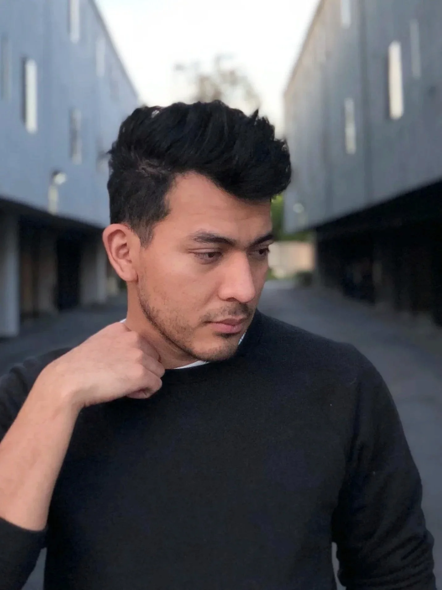 A young man with dark, styled hair, wearing a black shirt, poses in an alleyway between two modern buildings with metallic facades.