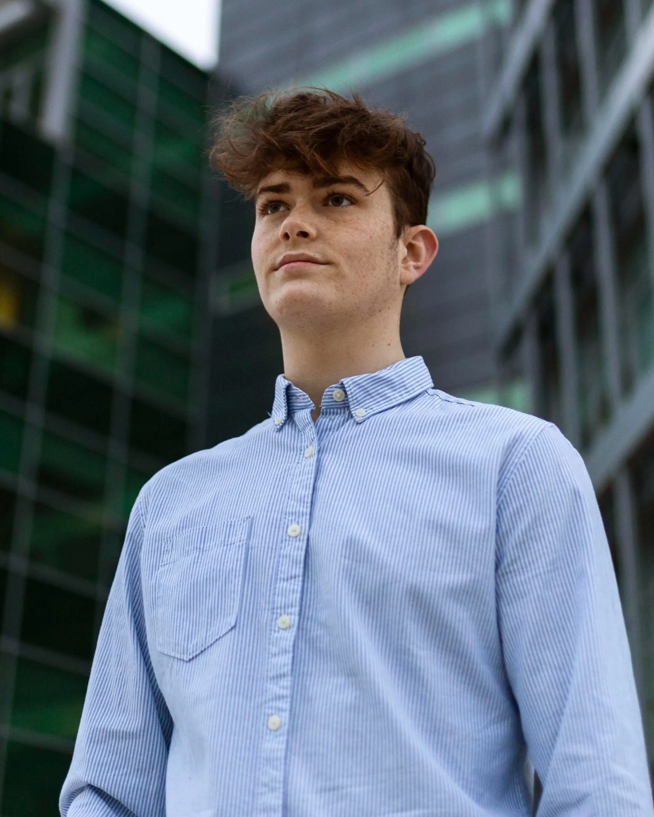 A young man with brown hair, wearing a blue and white striped button-up shirt, stands outdoors in front of modern glass buildings.