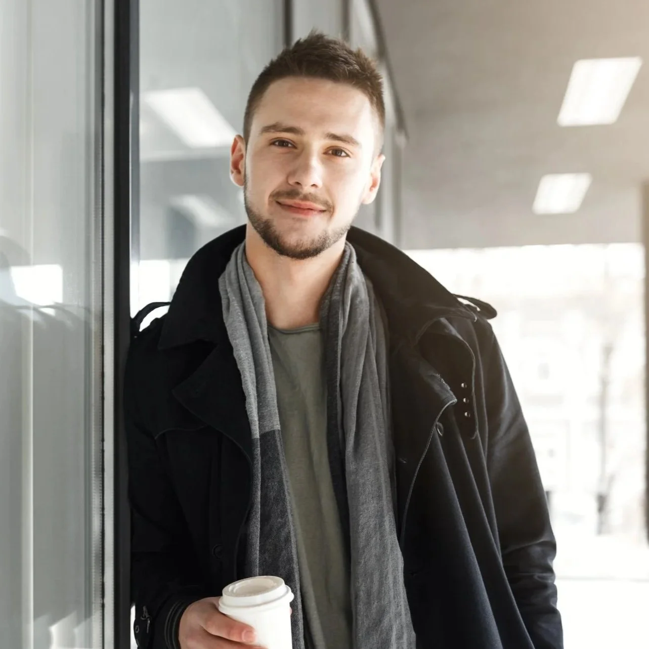 A young man with short brown hair and a beard holding a disposable coffee cup, leaning against a glass wall inside a modern building, wearing a black jacket and gray scarf.