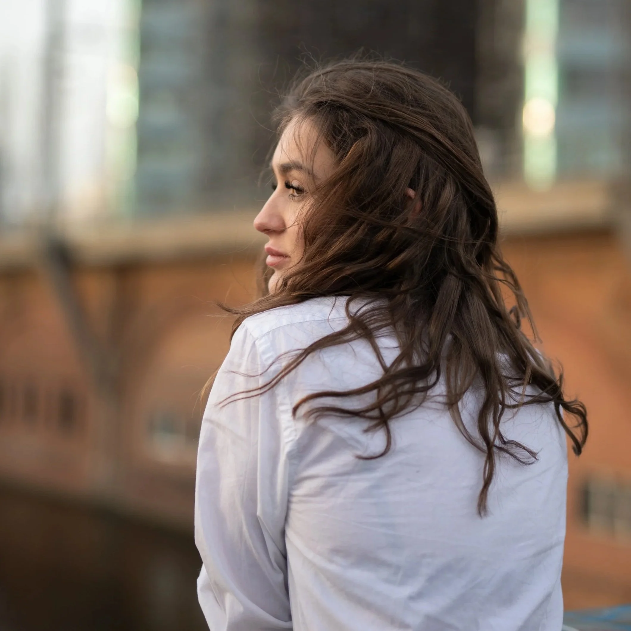 A woman with long, wavy brown hair wearing a white shirt, turned to the side with a contemplative expression in front of a blurred cityscape background.