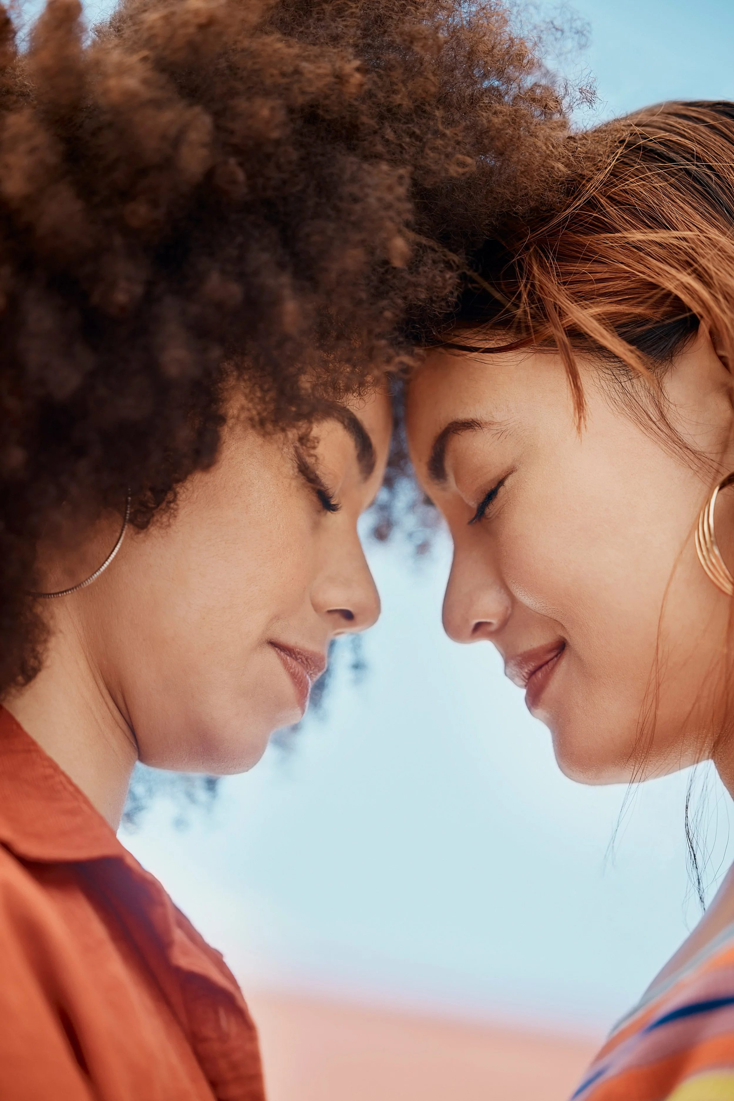 Two women with their foreheads touching, eyes closed and smiling softly, in a close-up portrait outdoors.