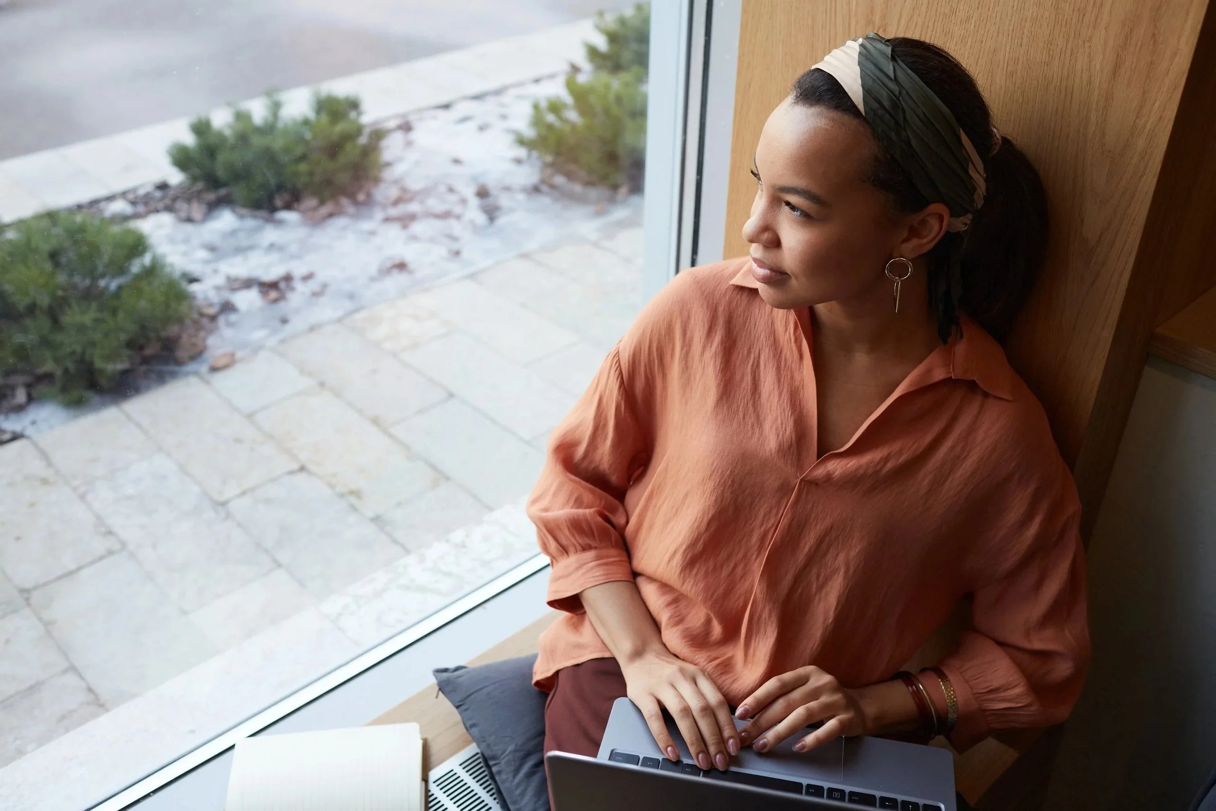 woman sitting by a window with a laptop on her lap, looking outside