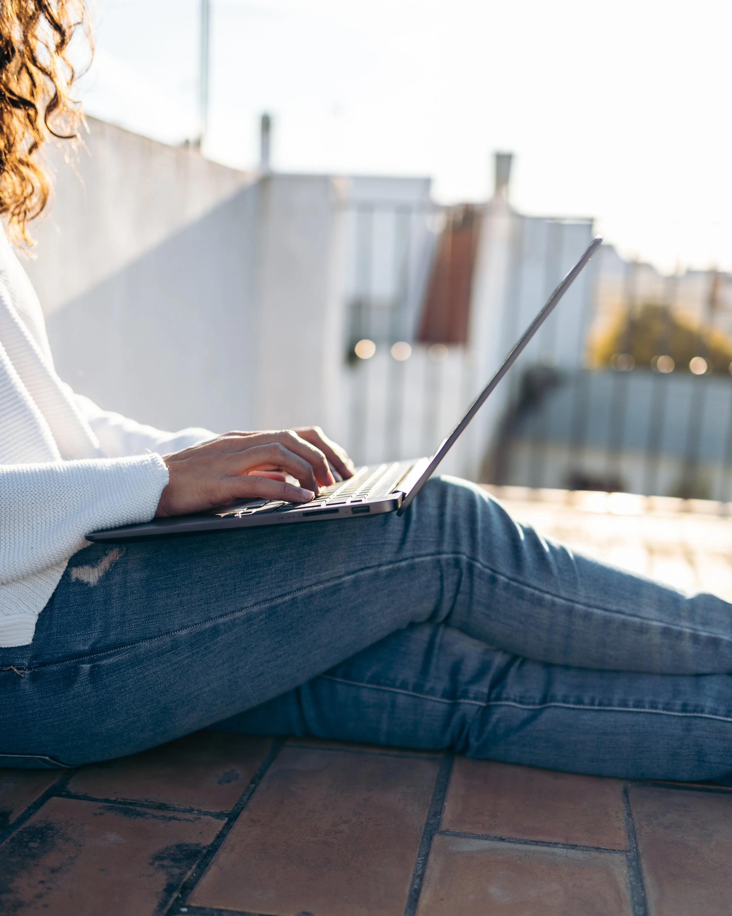 A person sitting outside on a tiled surface, using a laptop with sunlight in the background.