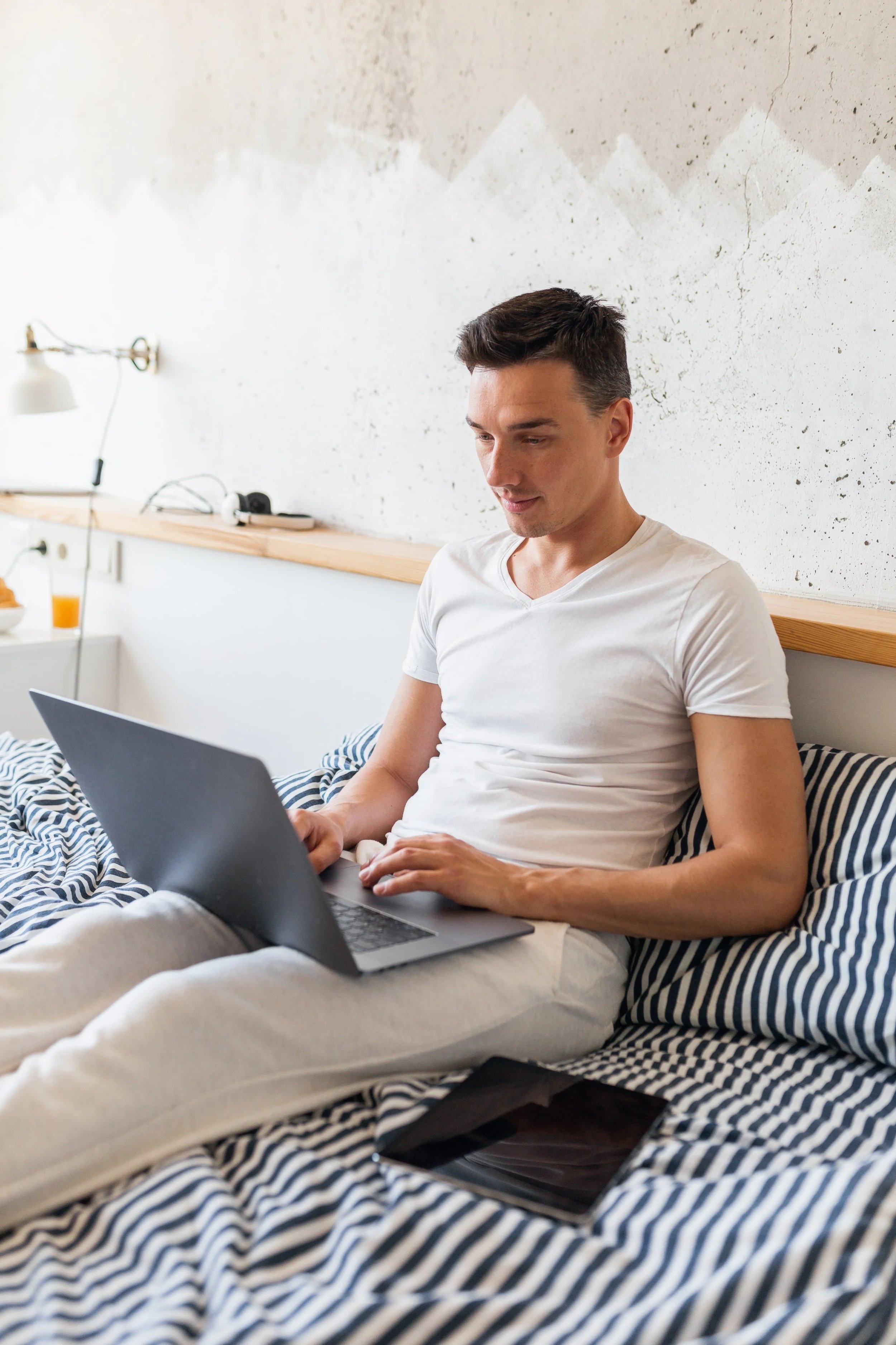 A young man with short dark hair sitting on a bed, using a silver laptop. He is wearing a white t-shirt and light-colored pants. There is a striped blanket on the bed and a tablet placed beside him. The background features a textured, off-white wall with wooden trim and some electronic devices on a shelf.