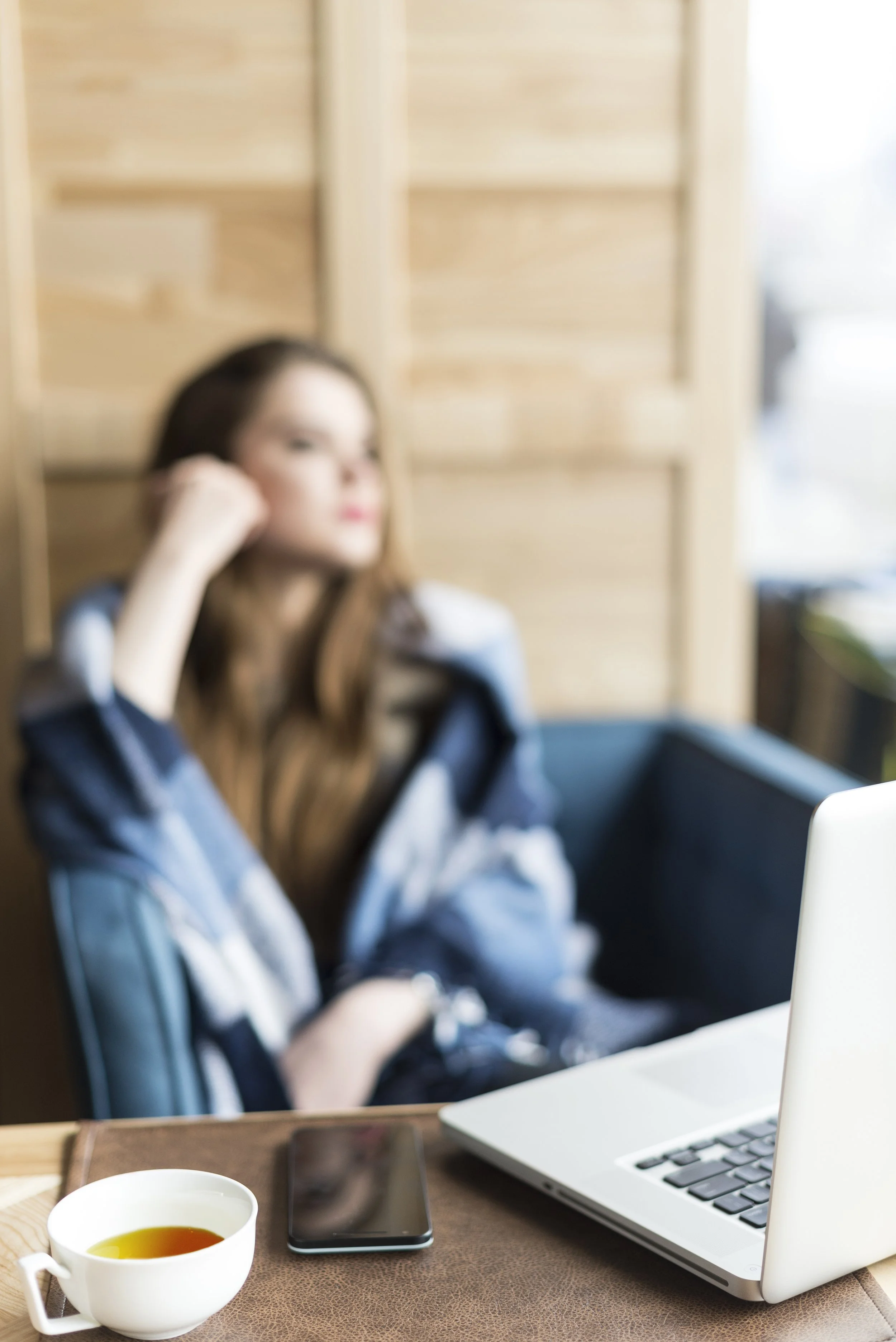 Blurry image of a woman sitting at a table with a laptop, smartphone, and a cup of tea or coffee in front of her, with a wooden partition in the background.