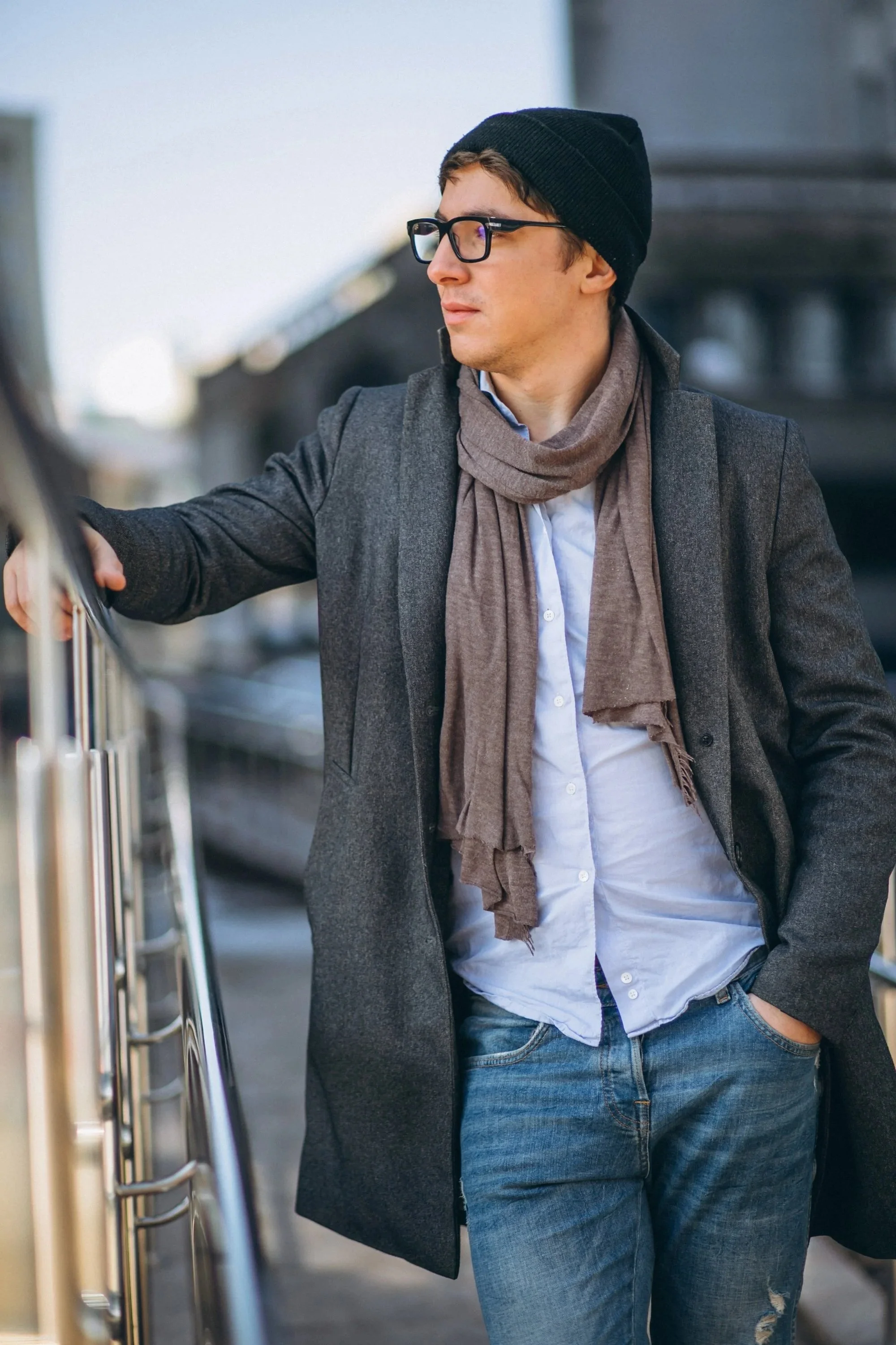A young man with glasses, a black beanie, and a gray coat stands outdoors, leaning on a metal railing with his right arm. He wears a white shirt, a brown scarf, and blue jeans, with his left hand in his pocket. The background shows a blurred cityscape.