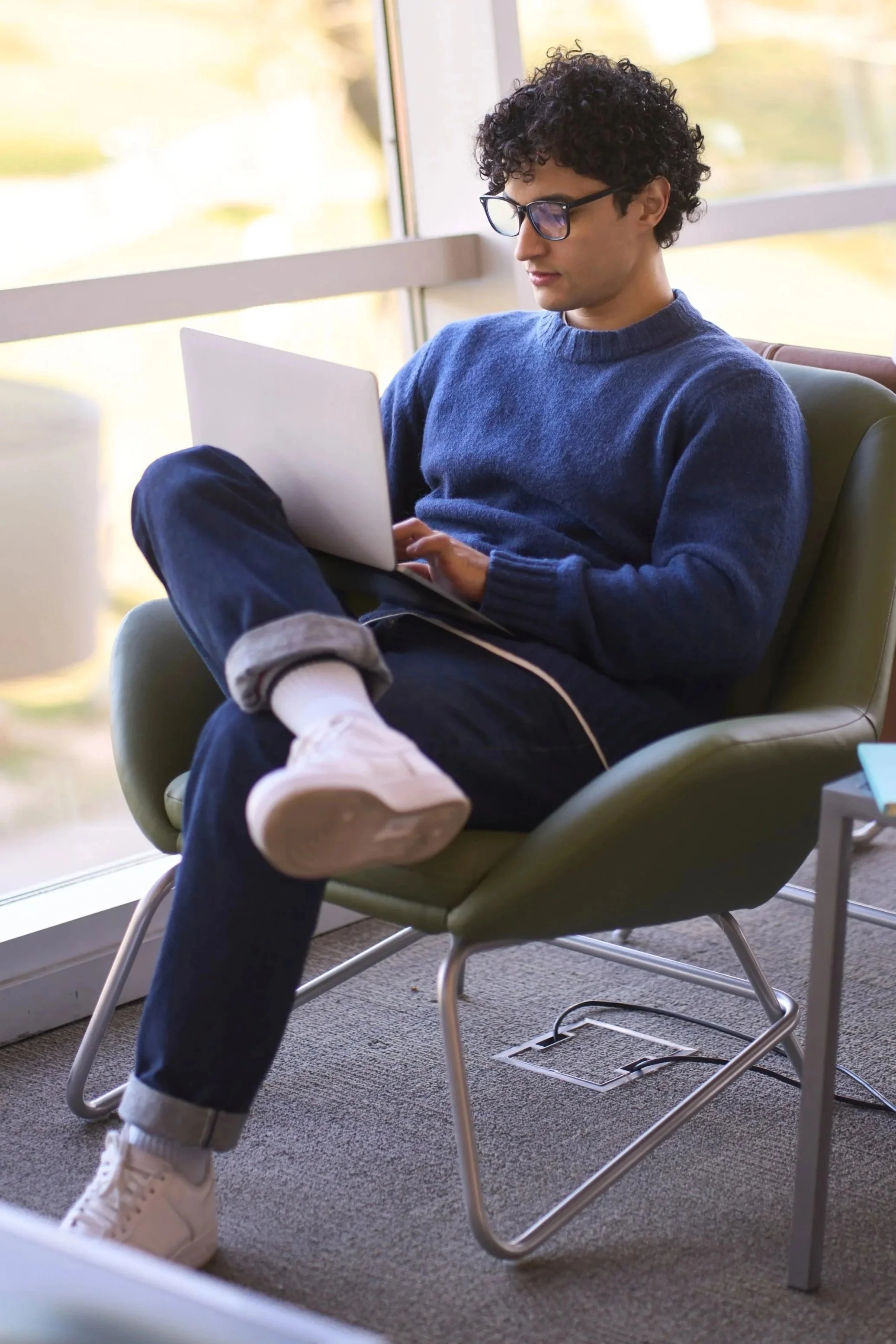 A young man sitting in a modern, green lounge chair by a large window, using a laptop. He is dressed casually in a blue sweater, white sneakers, and glasses, with one leg crossed over the other.
