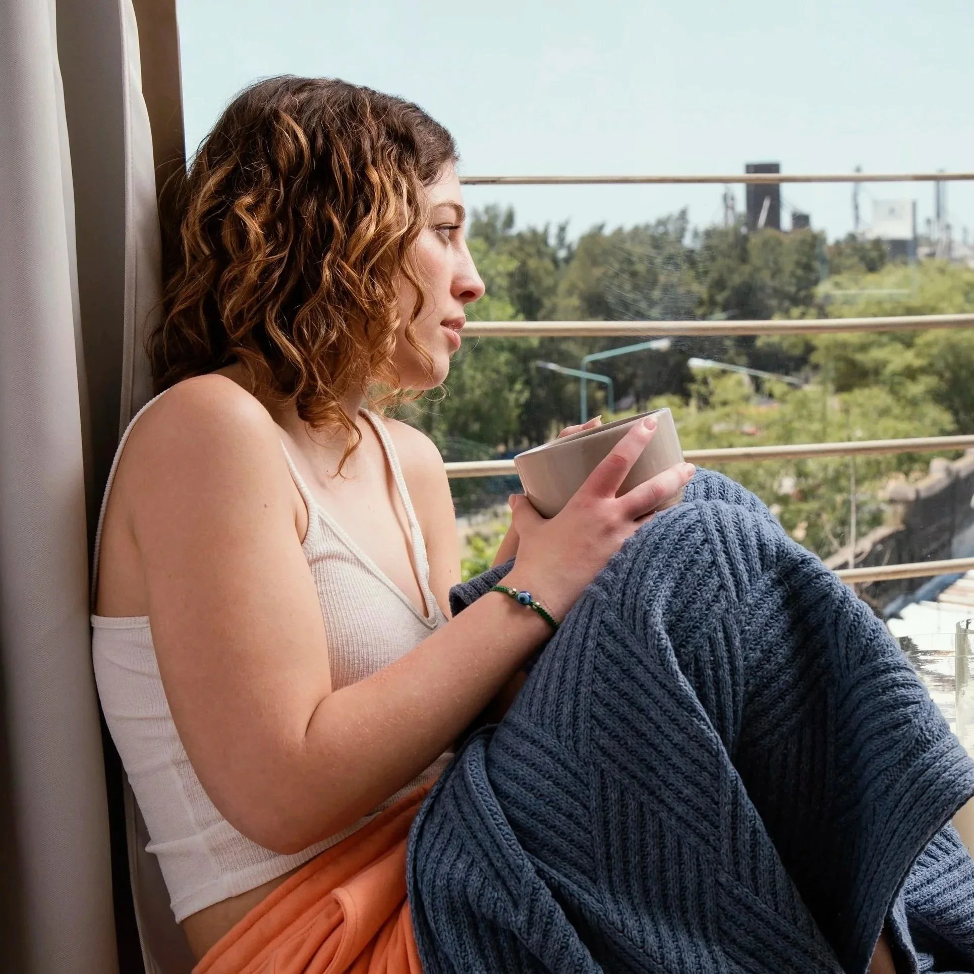 A woman with curly brown hair sitting by a window, holding a bowl and wearing a white tank top, blue knitted pants, and a bracelet, looking outside.