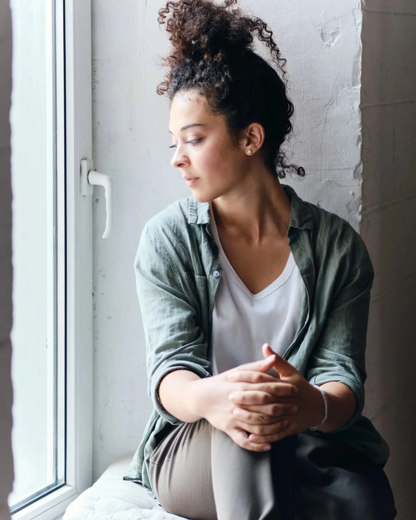 A woman with curly hair in a top knot, wearing a white shirt and green jacket, sitting by a window with her hands clasped and looking down thoughtfully.