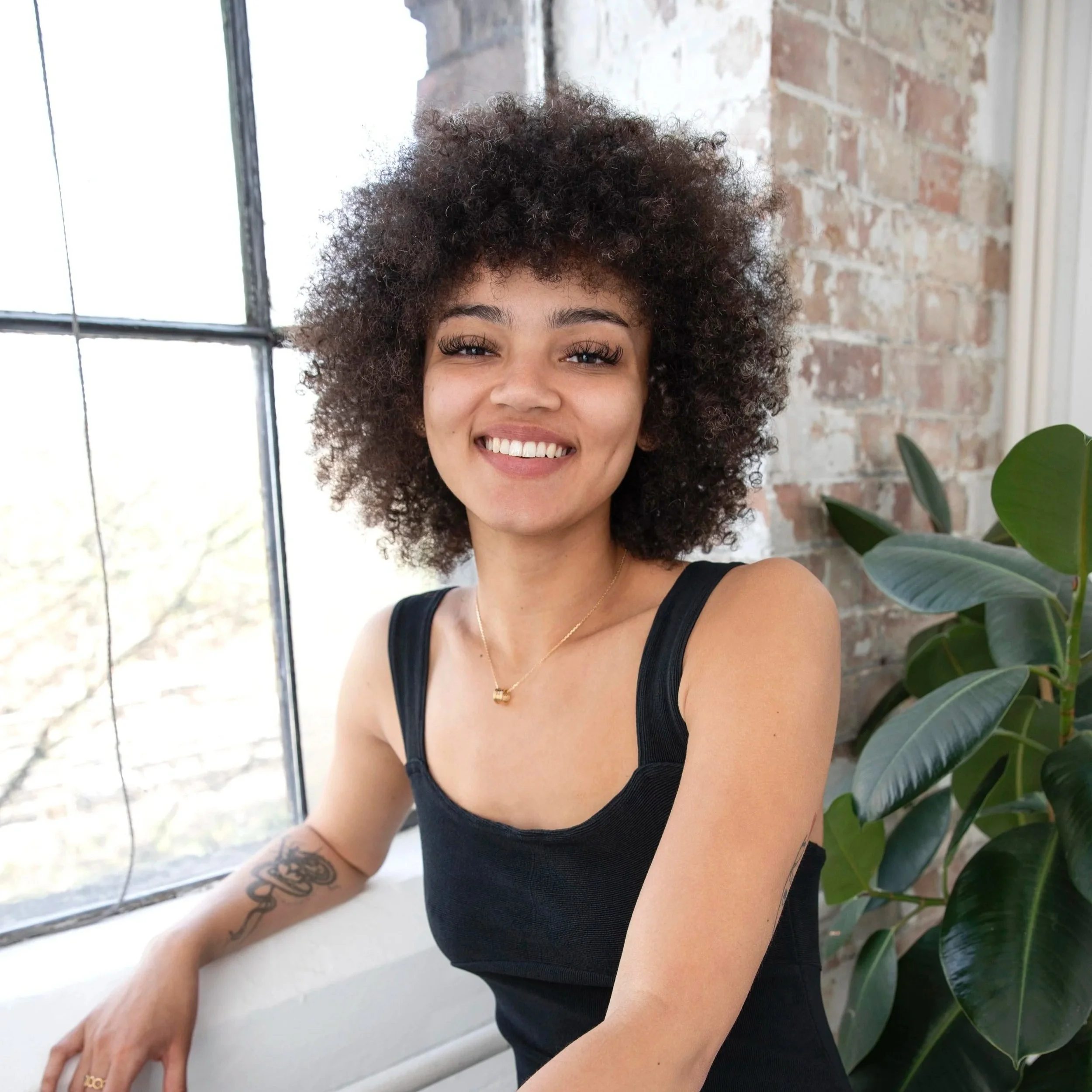 A young woman with curly hair, smiling with teeth showing, sitting near a large window with a brick wall behind her and a green plant beside her.