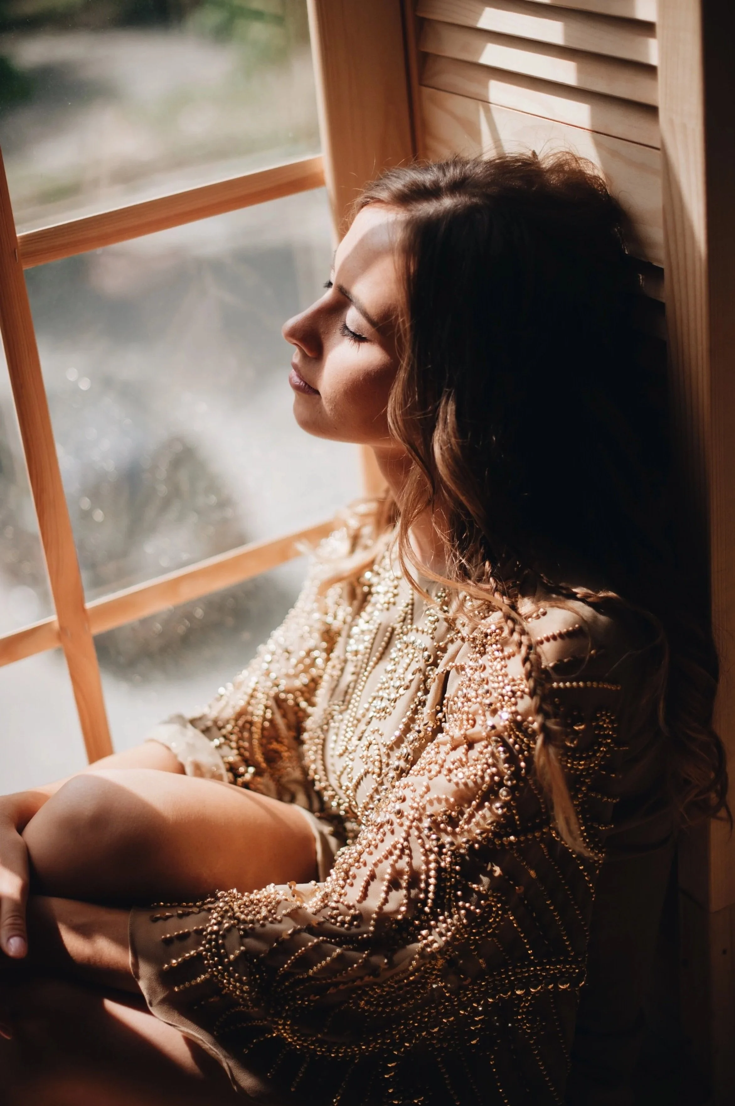 A woman with long dark hair and light skin sitting by a window with her eyes closed, wearing a beige dress adorned with pearls and beads, illuminated by sunlight.