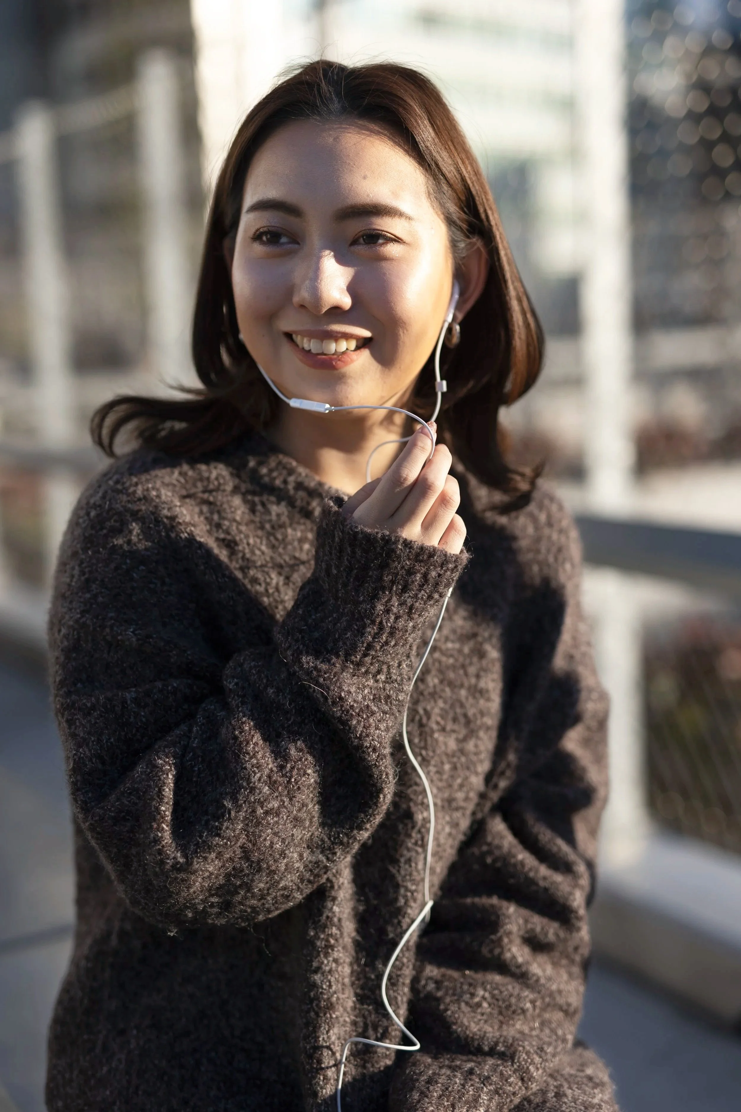 A woman smiling with earbuds, wearing a brown sweater, standing outdoors with a blurred background of a bridge and cityscape.