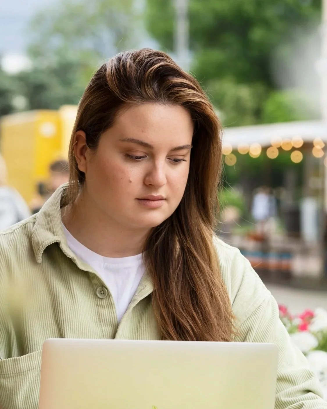 A young woman with long brown hair working on a laptop outdoors, with blurred trees and string lights in the background.