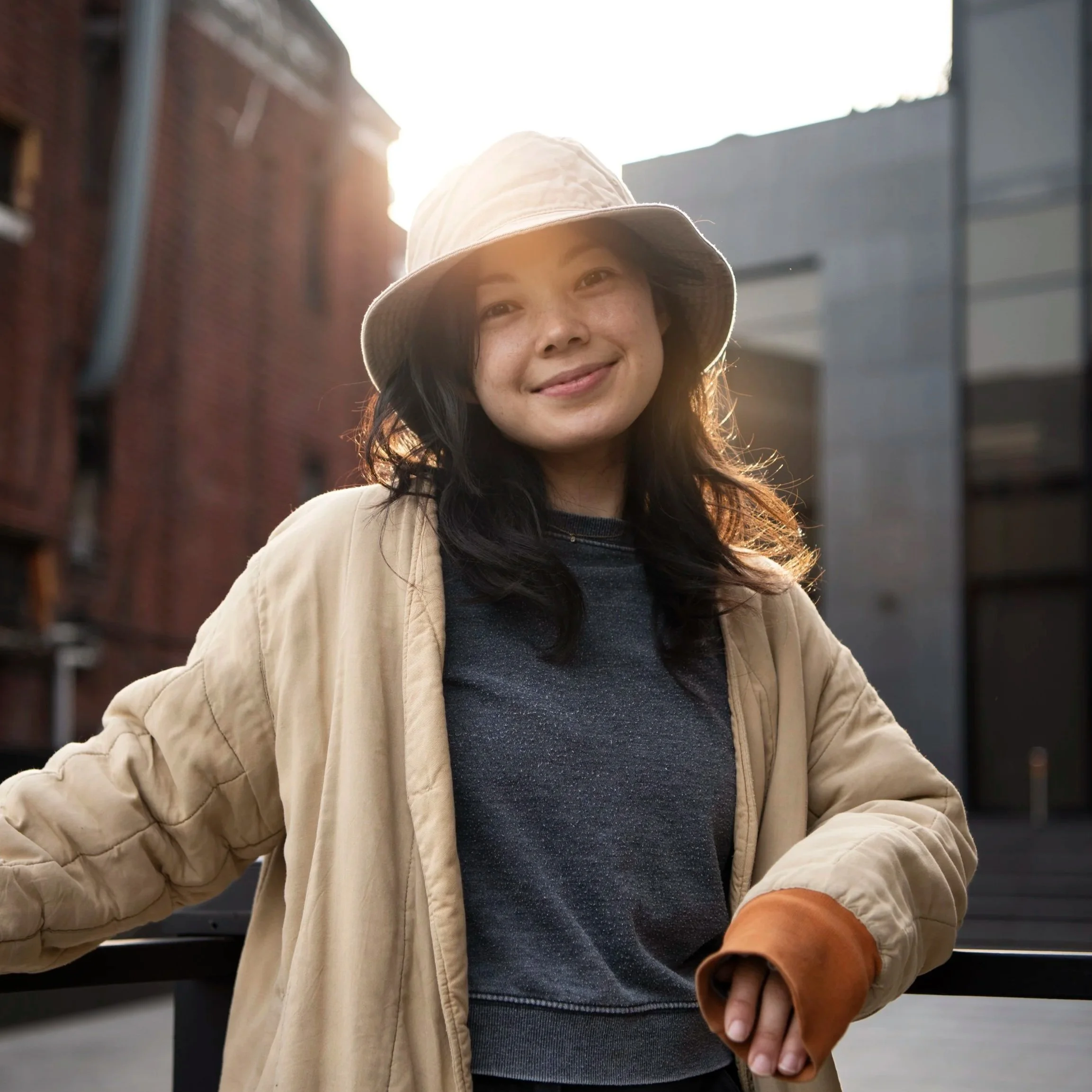 Woman wearing a beige jacket and a bucket hat, smiling in an urban setting at sunset.