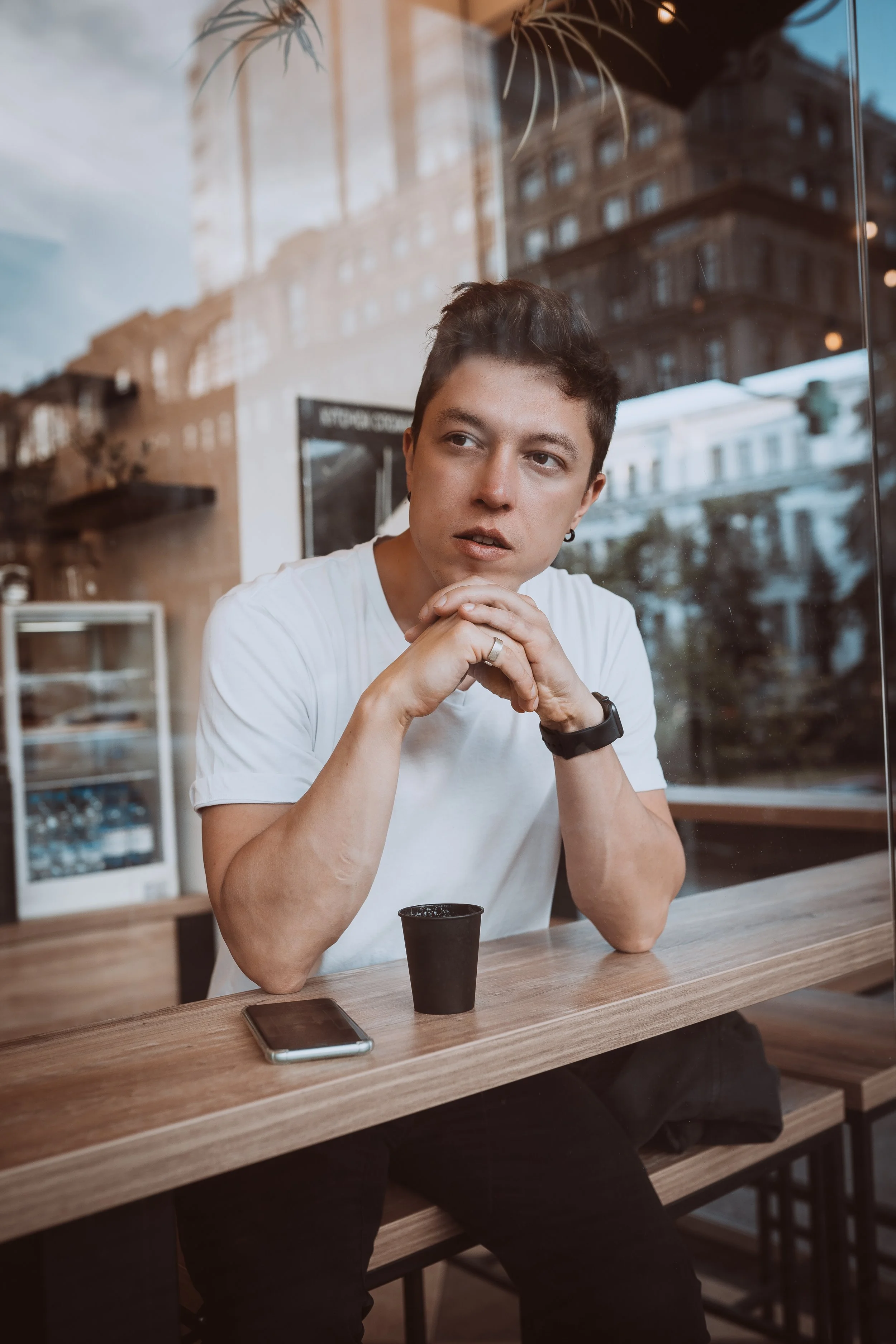 A young man sitting at a wooden table inside a coffee shop, looking out the window with a contemplative expression. There is a smartphone and a small black cup on the table.