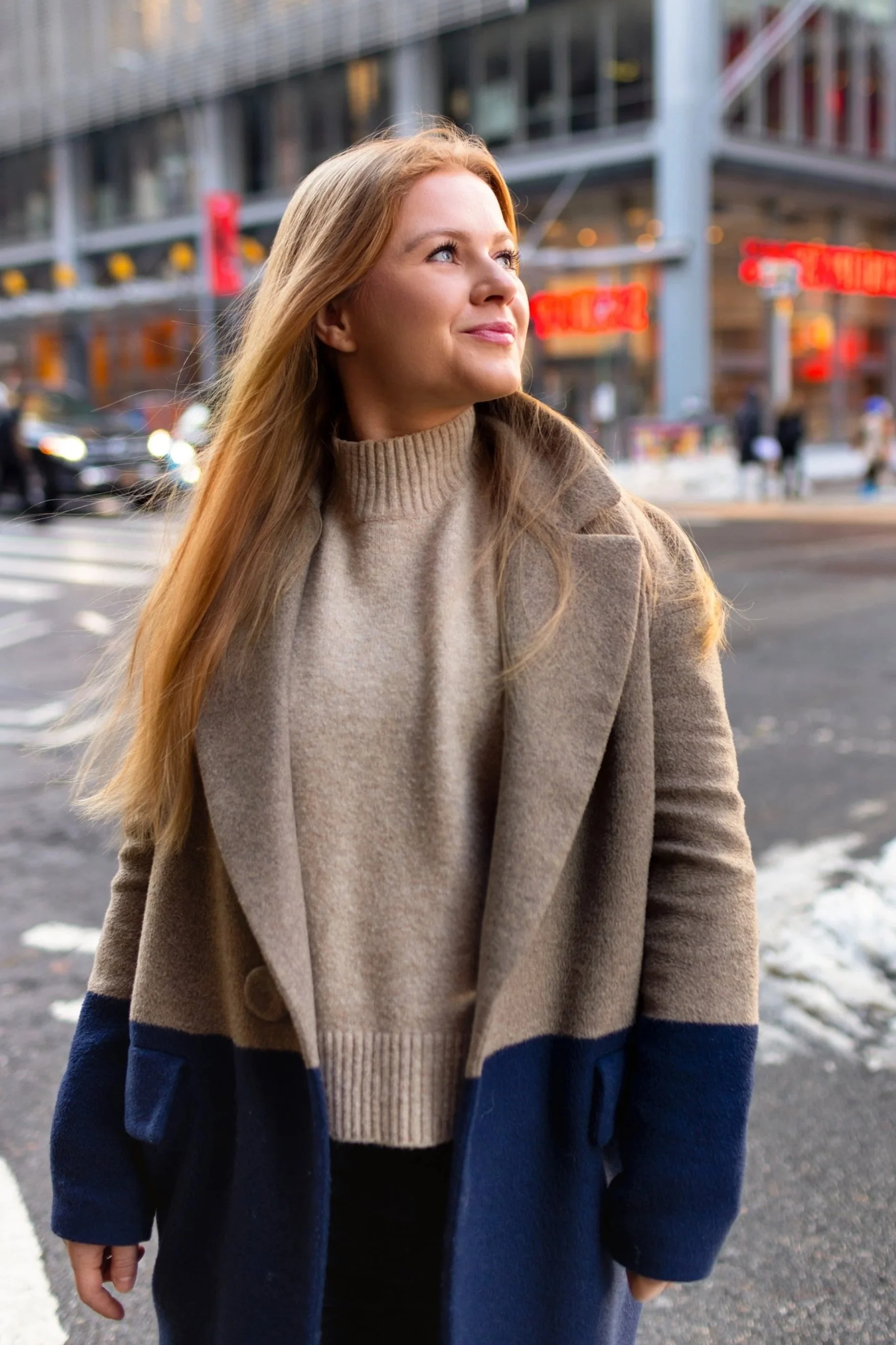 A woman with long blonde hair wearing a beige turtleneck sweater and a beige and navy coat standing at a city street corner in the evening.