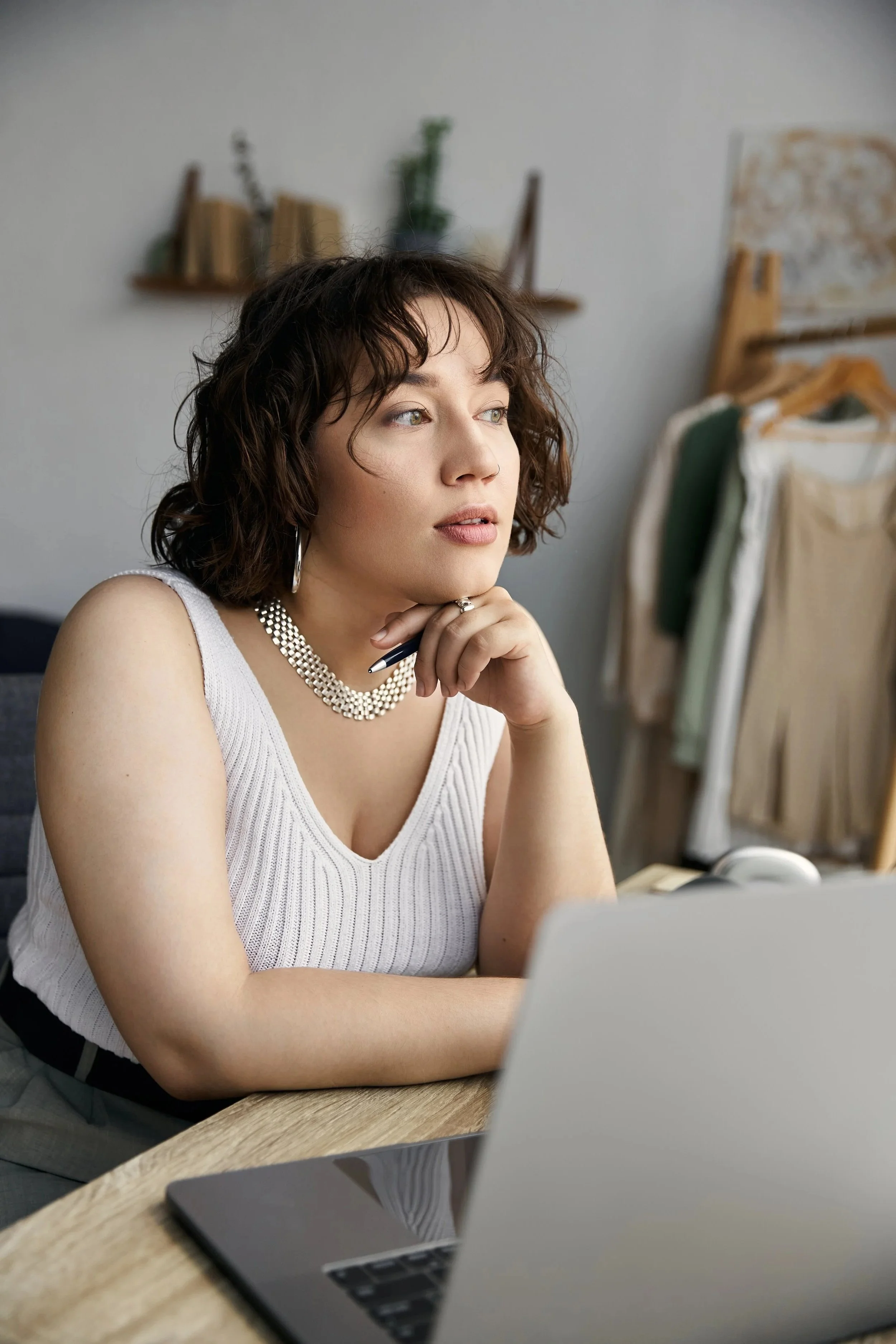 A woman with curly brown hair, wearing a white sleeveless top, silver earrings, and a pearl necklace, is sitting at a wooden desk with a laptop in front of her. She appears to be deep in thought, with her chin resting on her hand and a pen touching her face. In the background, there is a clothing rack with various garments and a shelf with books and decorative items.