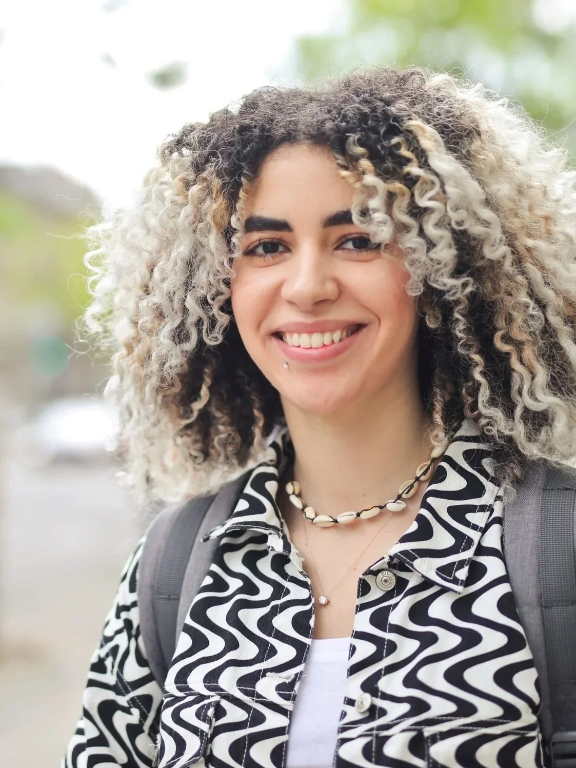 A young woman with curly blonde and black hair, smiling, wearing a patterned black and white jacket, necklaces, and carrying a backpack outdoors.