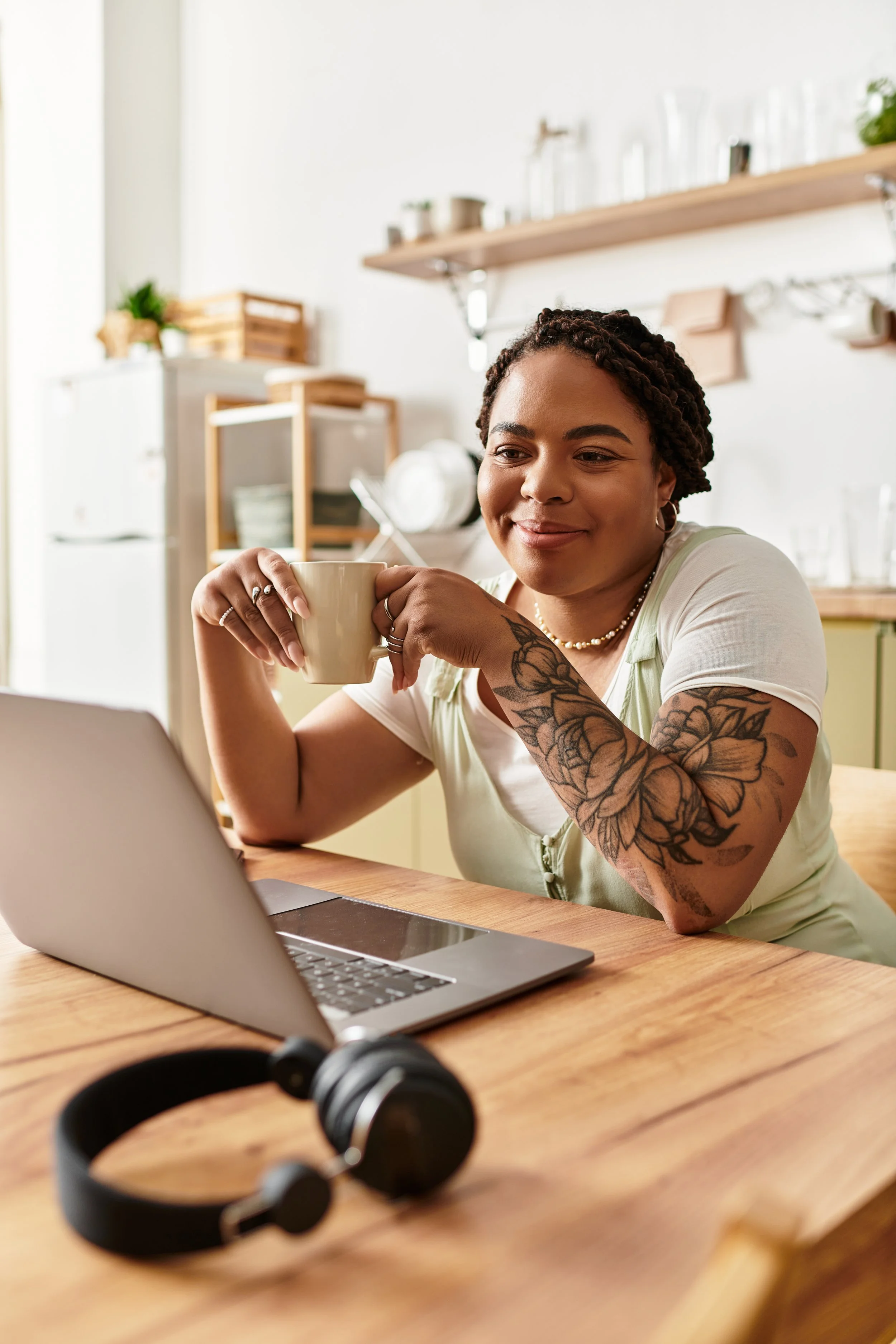 A woman with tattoos on her arms and wearing jewelry is sitting at a wooden table in a bright kitchen, holding a beige mug, looking at her laptop screen, with black headphones on the table in front of her.