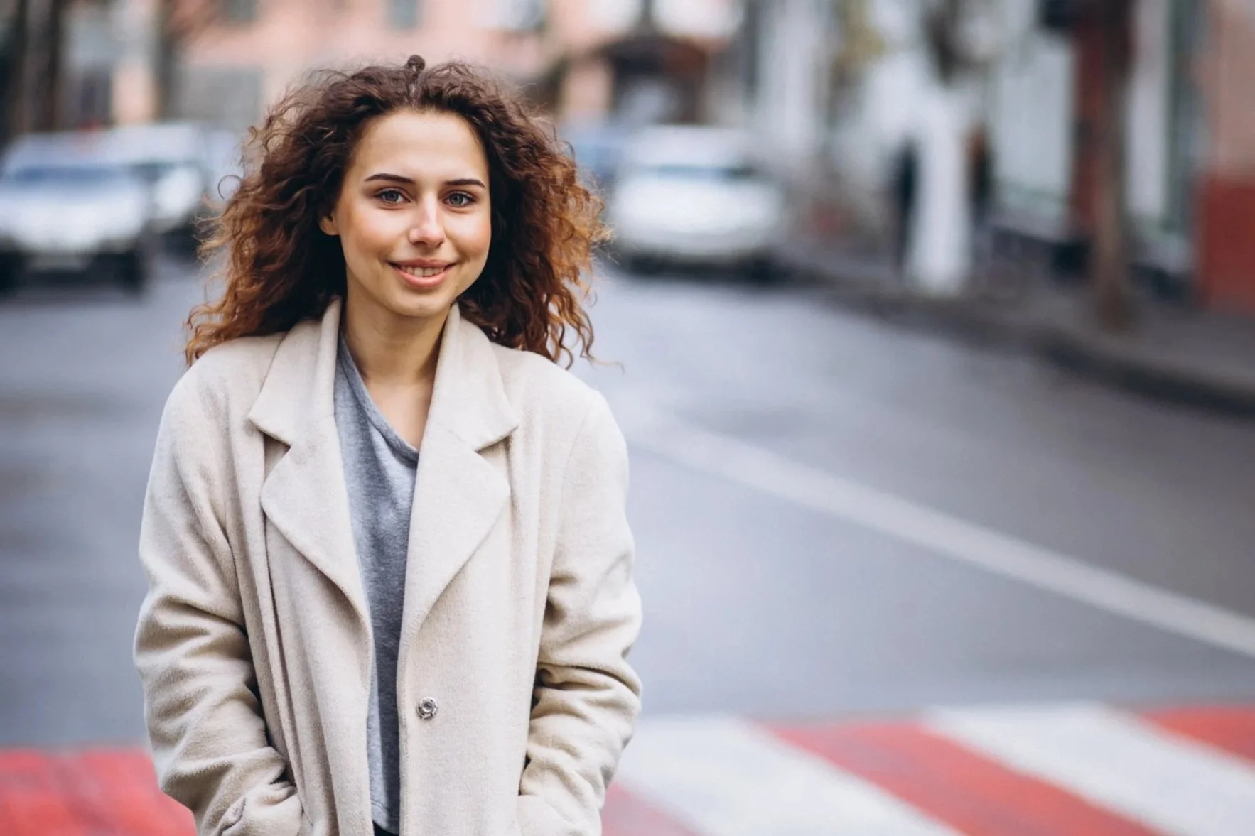 Young woman with curly hair wearing a beige coat standing on a city street.