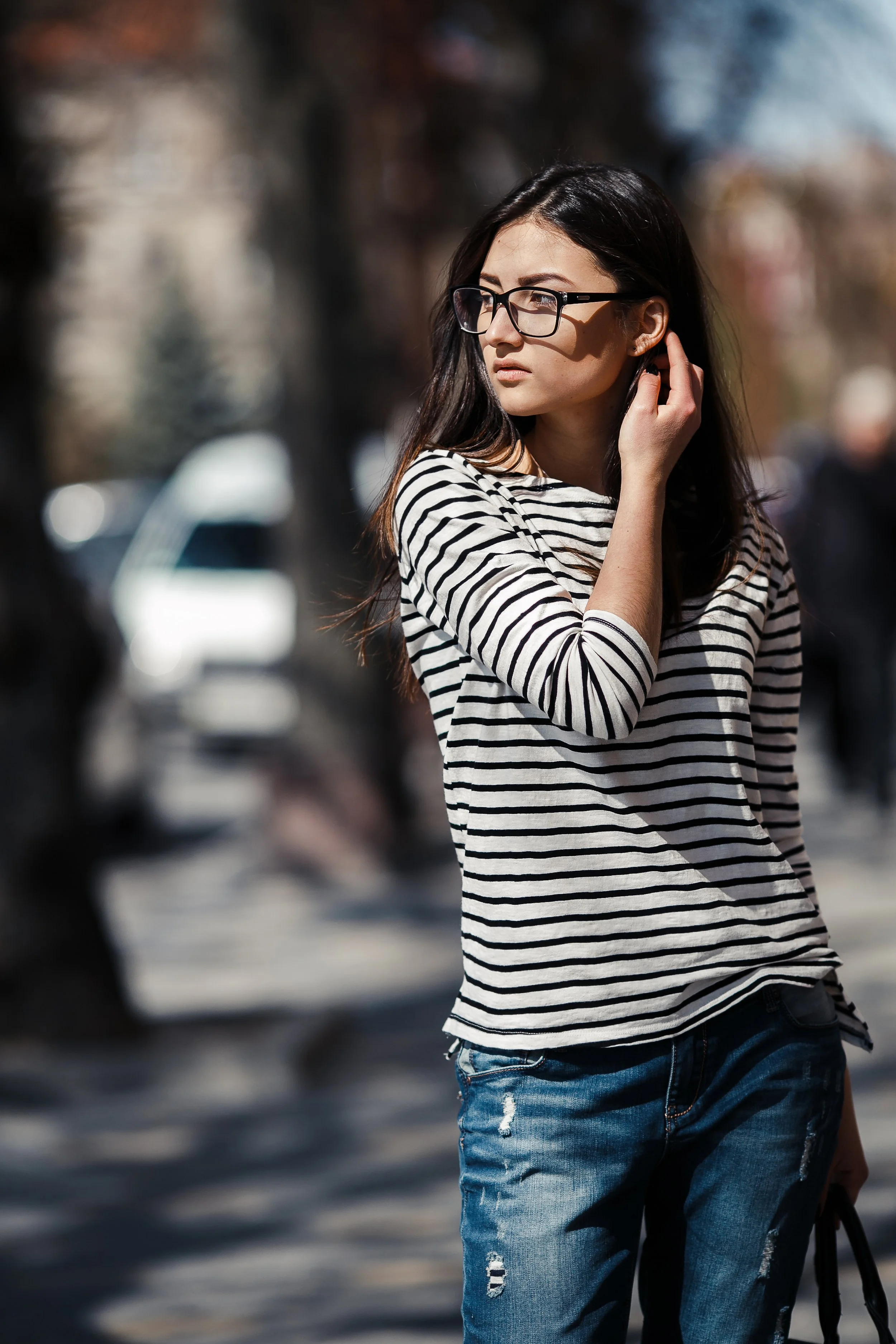 A woman with long dark hair, wearing glasses, a striped shirt, and distressed jeans, standing outdoors on a sunny day.