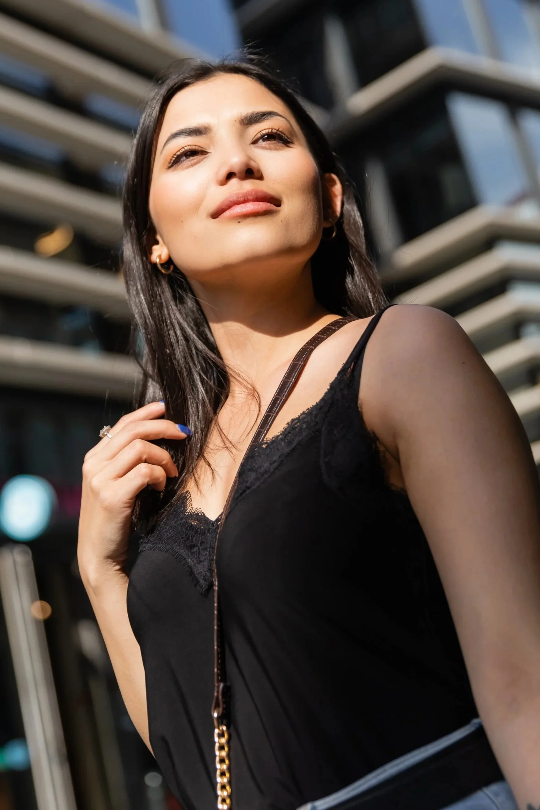 A young woman with dark hair, wearing a black sleeveless top, stands outdoors in sunlight with modern office buildings in the background.