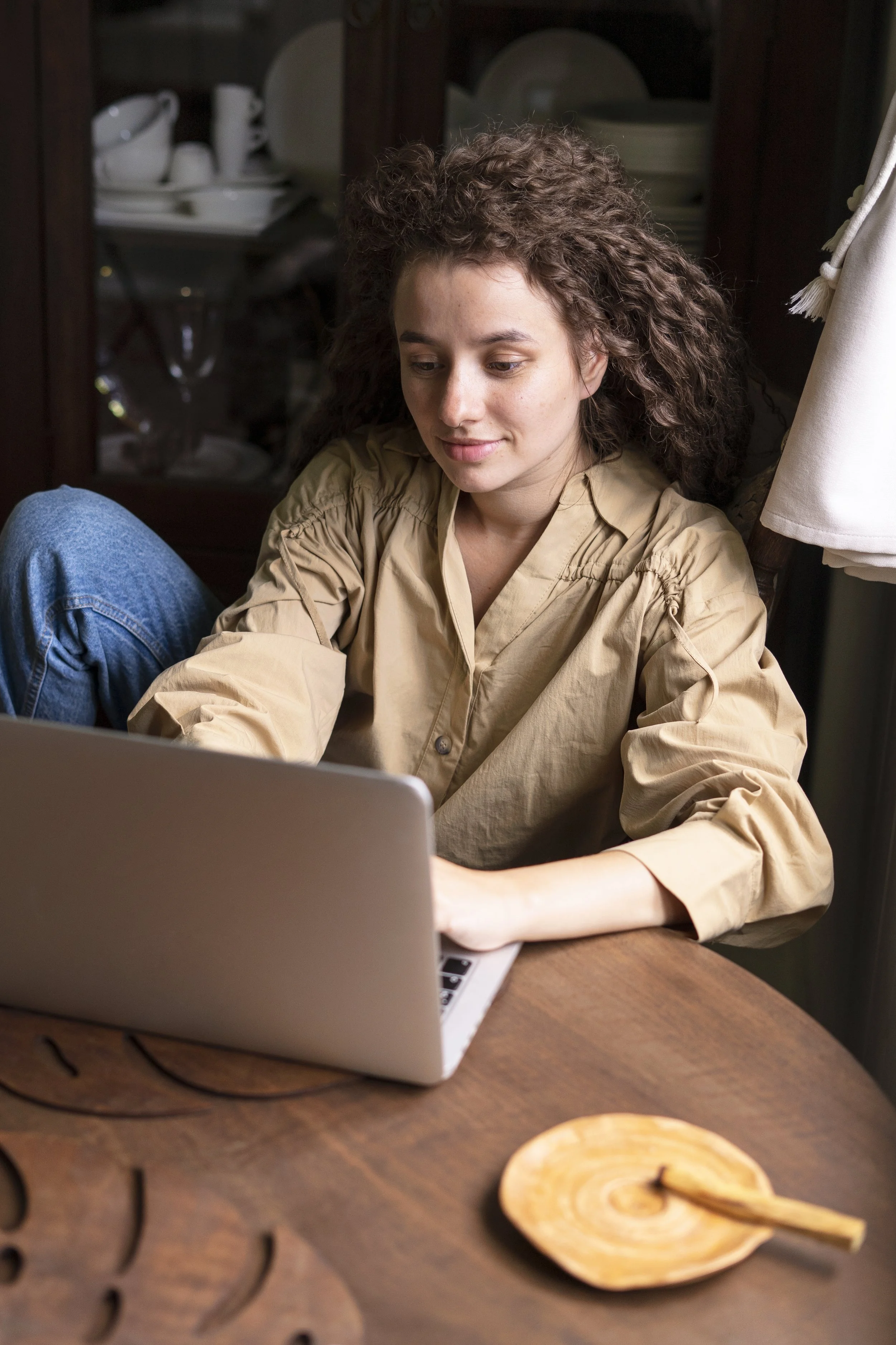 A young woman with curly hair sitting at a wooden table working on a laptop.