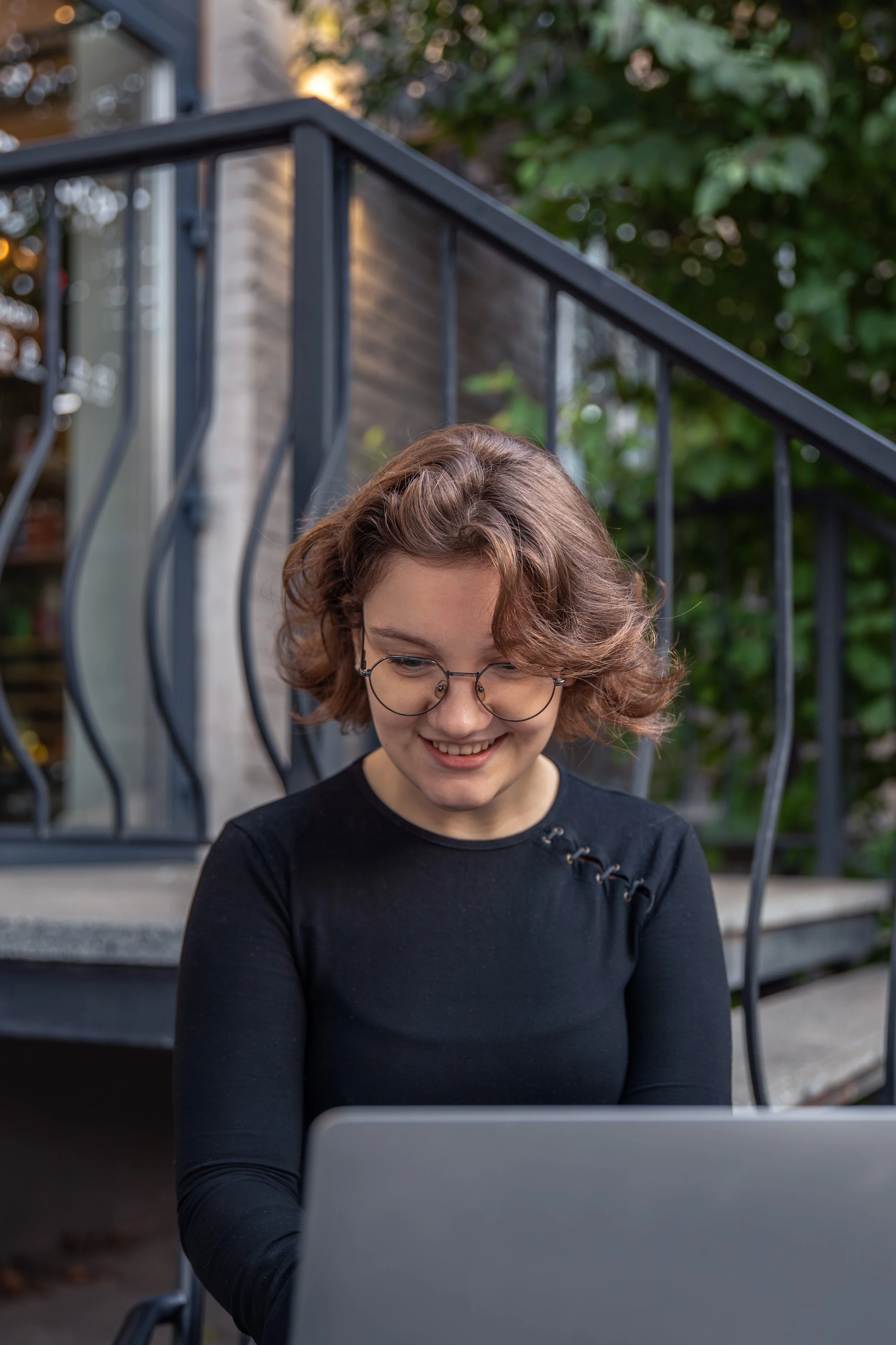 A young woman with curly short hair, glasses, and a black long-sleeve shirt, sitting outside on the ground near stairs, working on a laptop, smiling, with a background of greenery and a house with a metal railing.