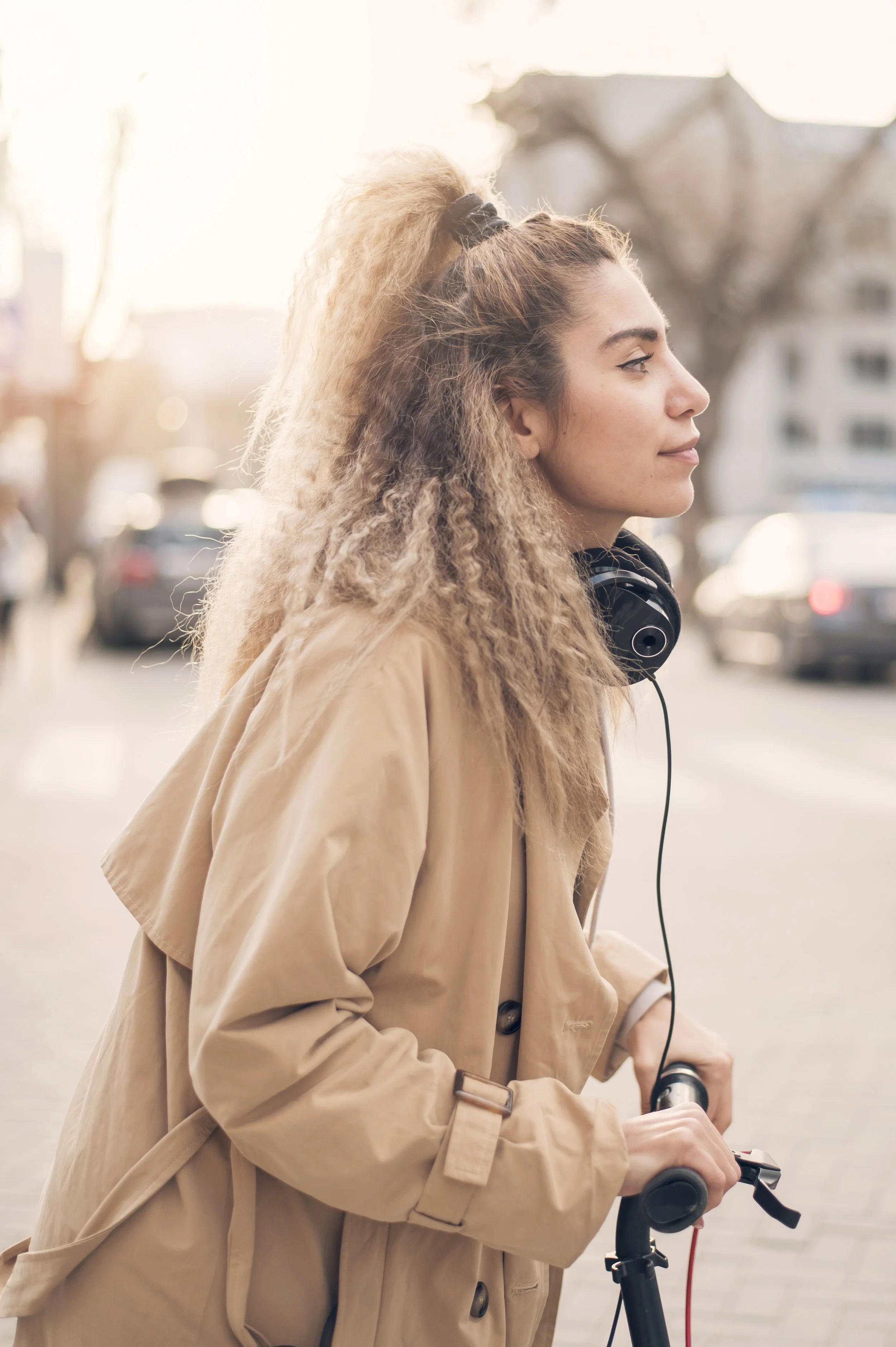 A young woman with curly blonde hair riding an electric scooter on a city street at sunset, wearing a beige trench coat and black headphones around her neck.