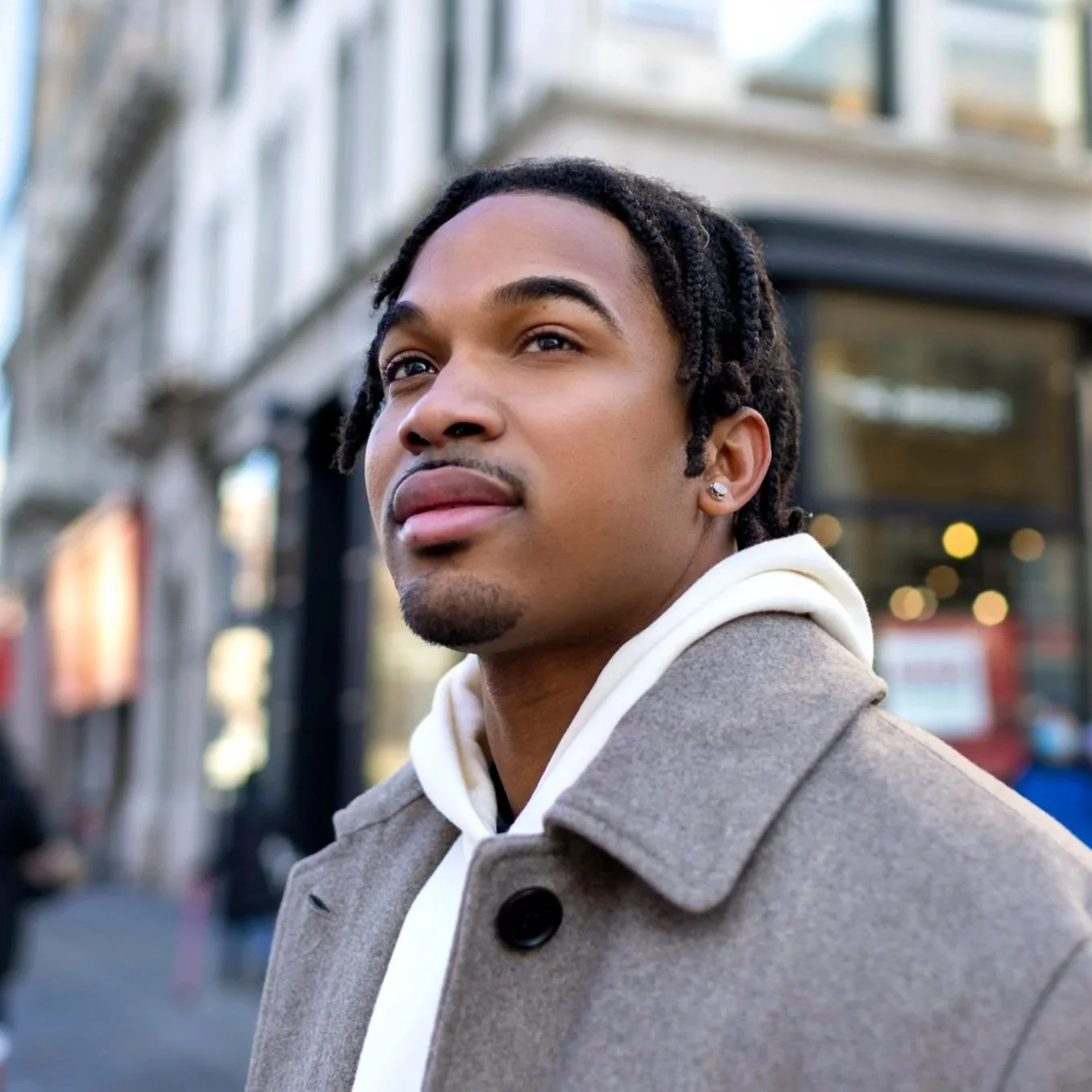 Young man with dreadlocks wearing a beige coat and hoodie on city street.