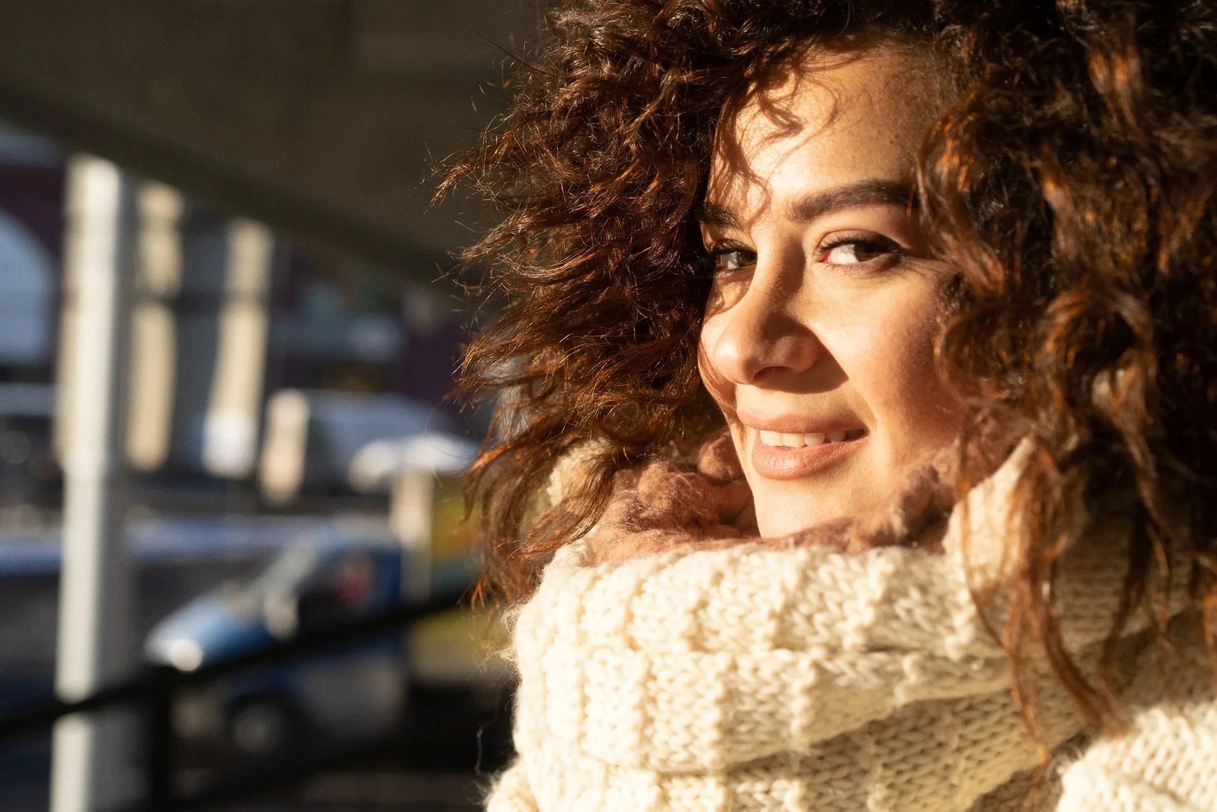 A woman with curly brown hair smiling in an outdoor setting, wearing a cream-colored knit scarf and sweater, with a blurry parking lot in the background.