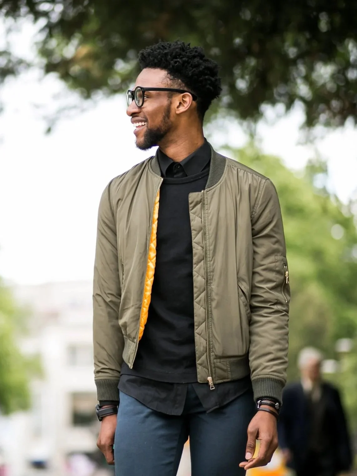 A young man with glasses and a beard, smiling and standing outdoors with trees in the background. He is dressed in a black shirt, paired with a green bomber jacket, and has a black bracelet on his wrist.