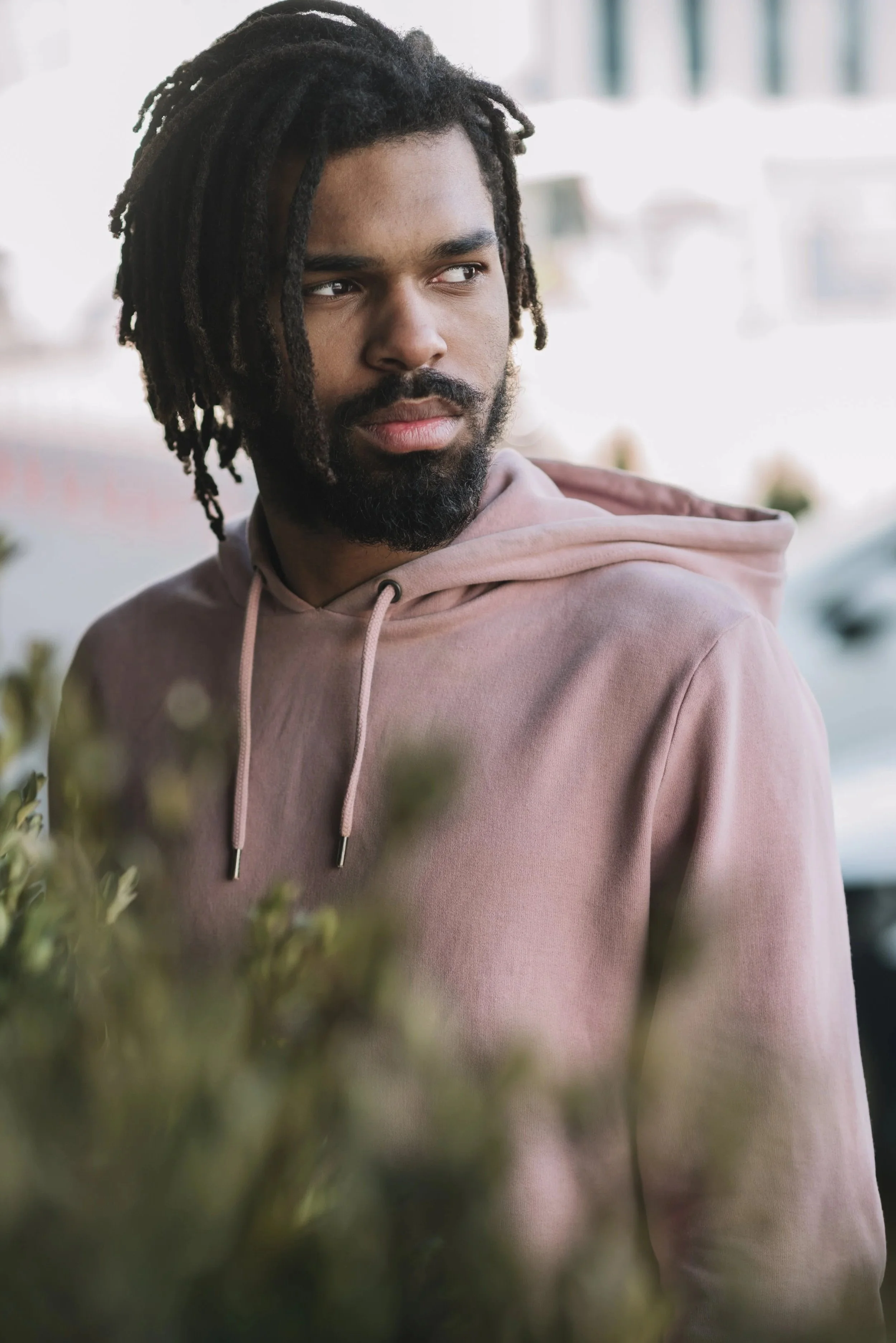 A young man with dreadlocks and a beard looking to the side, wearing a pink hoodie, outdoors with blurred background.
