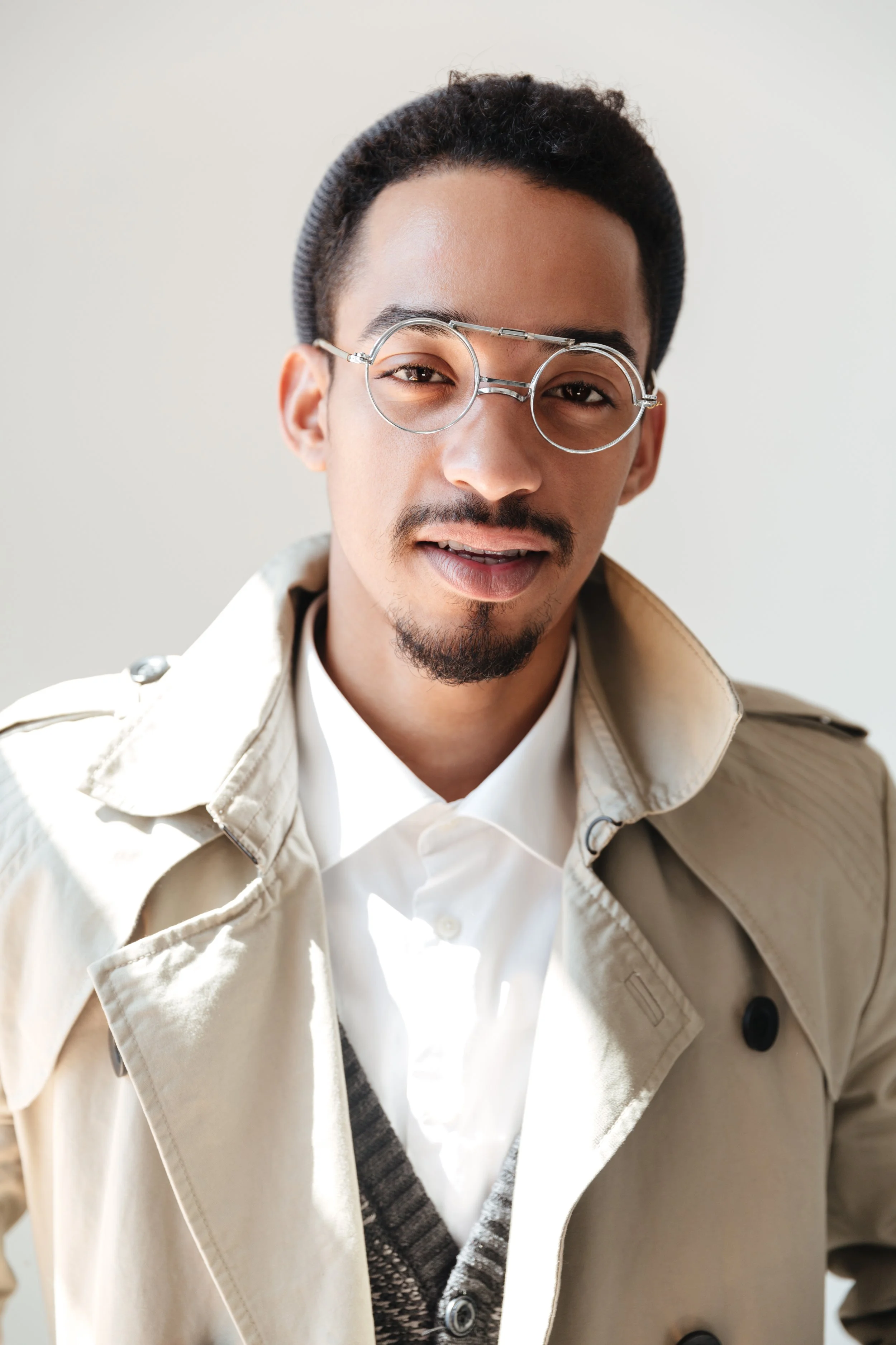 A young man with glasses, a goatee, and wearing a beige trench coat over a gray sweater and white shirt, smiling at the camera.