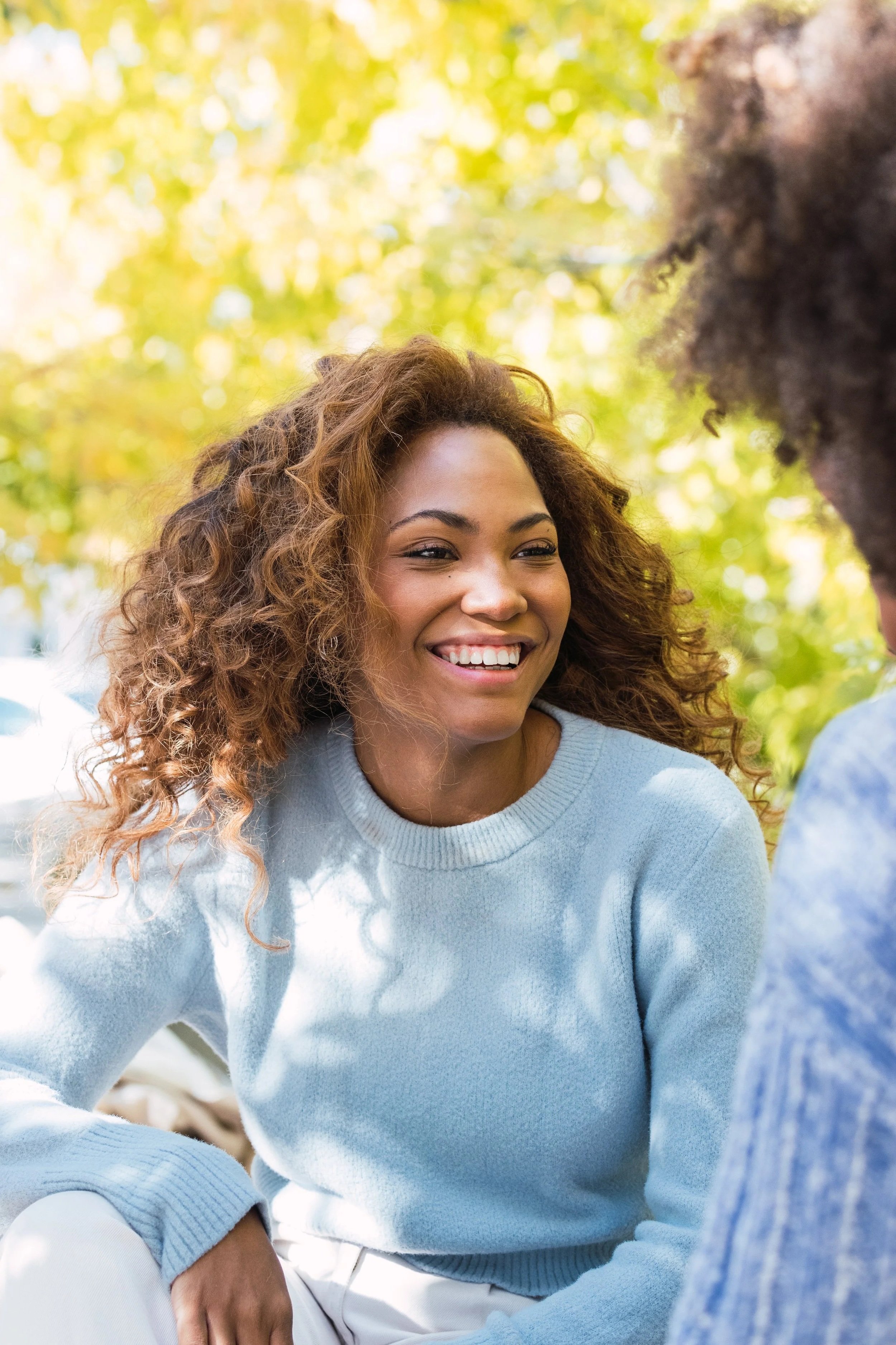 A woman with curly hair smiling and talking with a person with an afro hairstyle outdoors in a park with yellow and green trees.