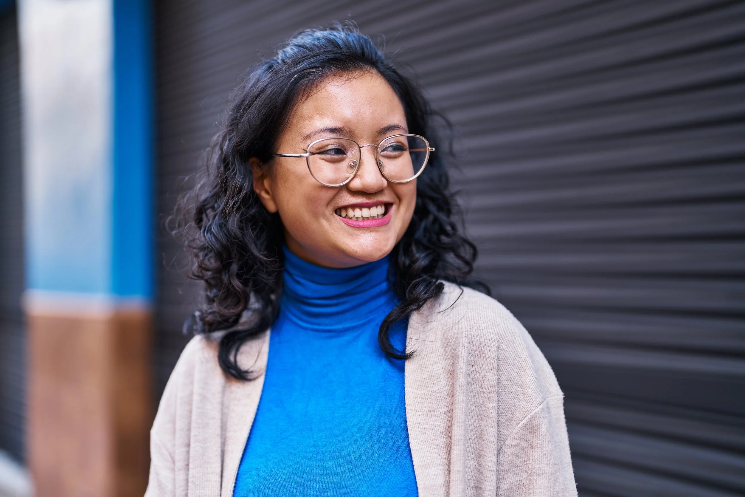 A woman with black curly hair, wearing glasses, a bright blue turtleneck, and a beige cardigan, smiling outdoors near a dark, ribbed metal wall.