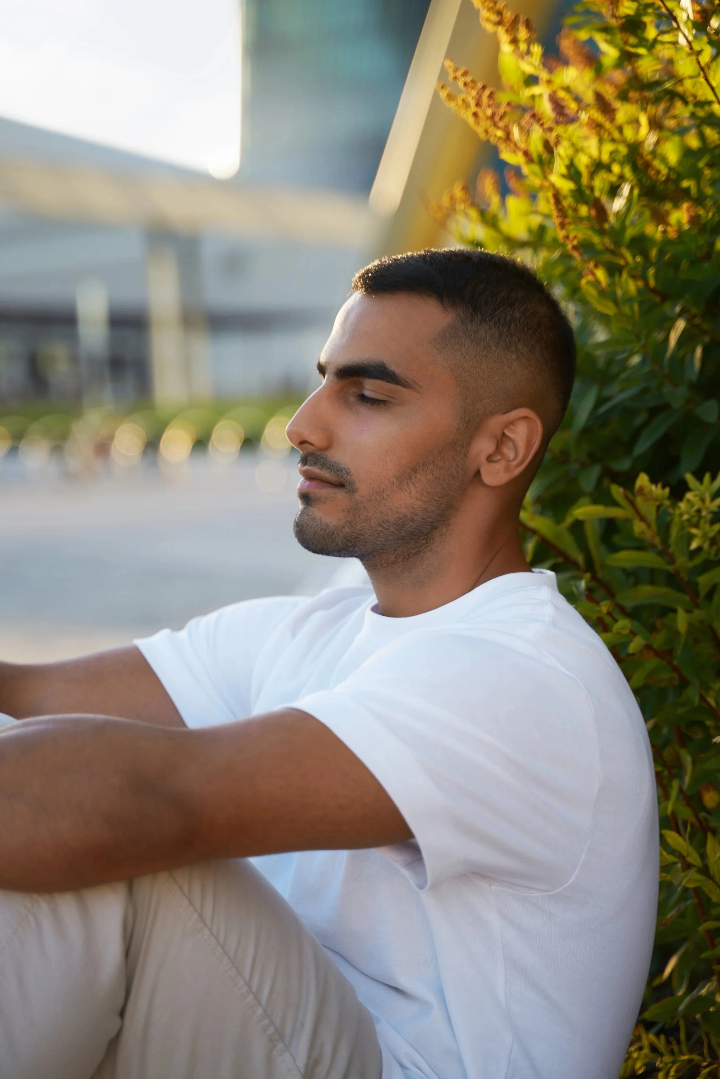 A young man with short hair and a beard, sitting outdoors with eyes closed, leaning against a greenery wall, during golden hour.