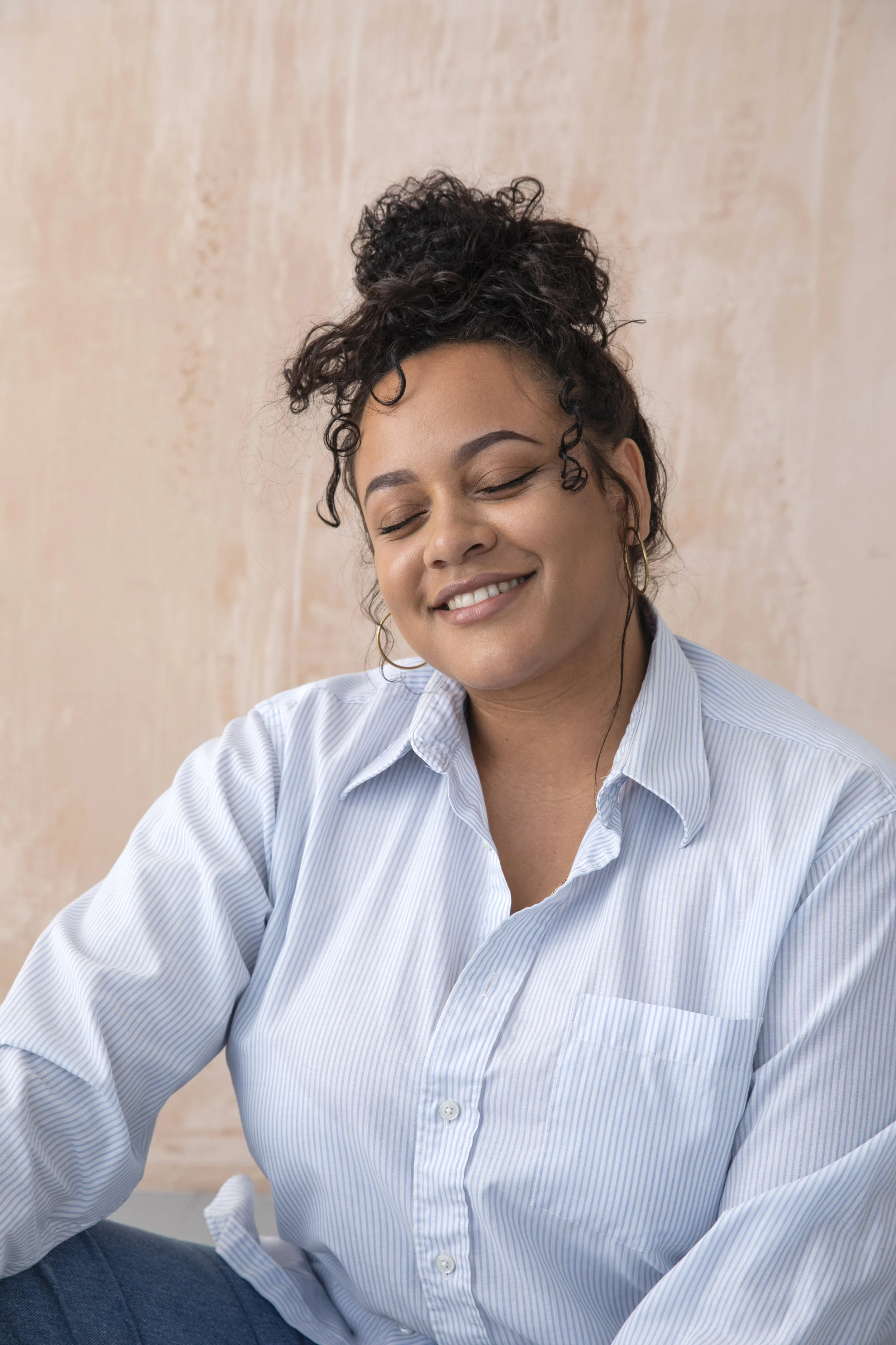 A woman with curly dark hair in a bun, wearing a light blue striped shirt and hoop earrings, smiling with eyes closed.