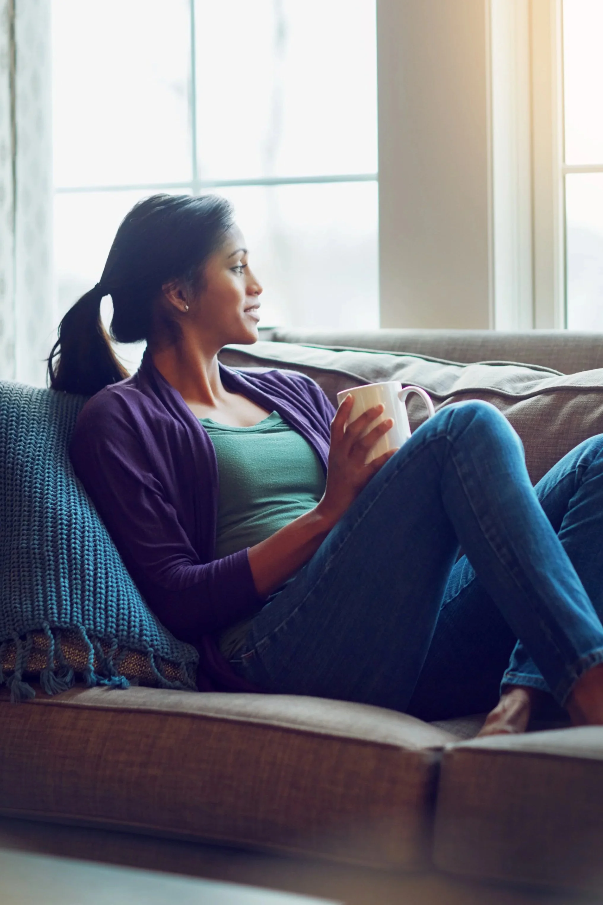 Woman sitting on a couch holding a mug, looking relaxed near a window.