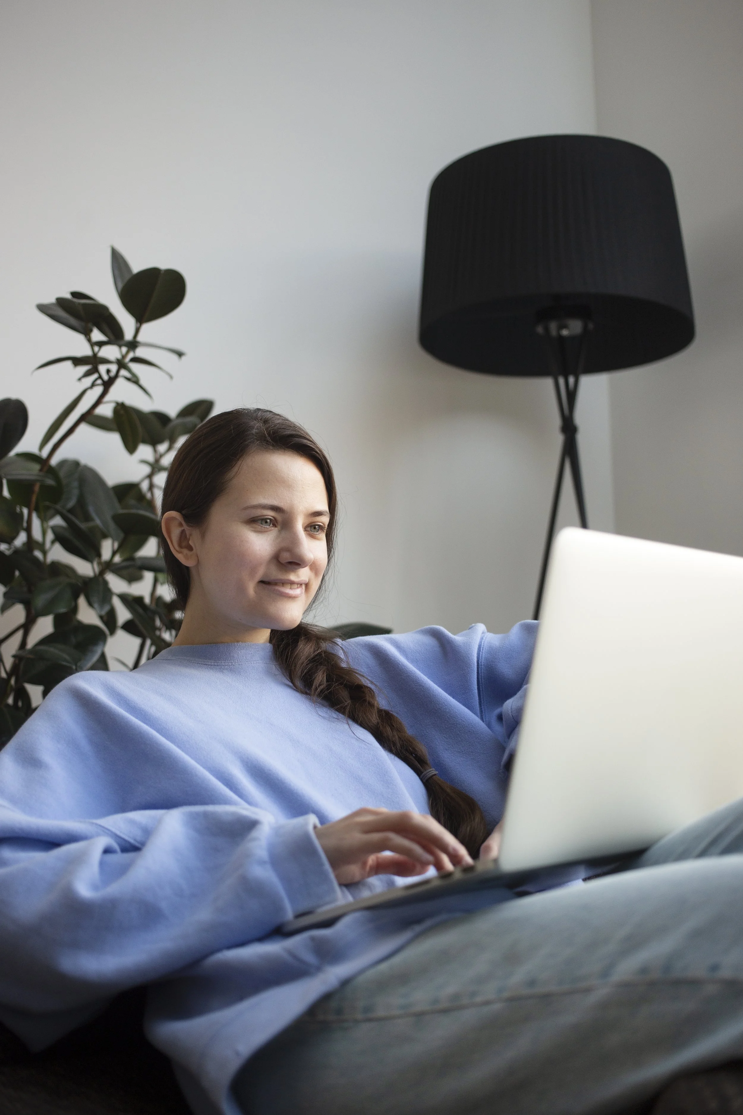 A woman with long brown hair in a braid, wearing a light blue sweatshirt, sitting on a couch, using a silver laptop, with a black floor lamp and a green plant in the background.
