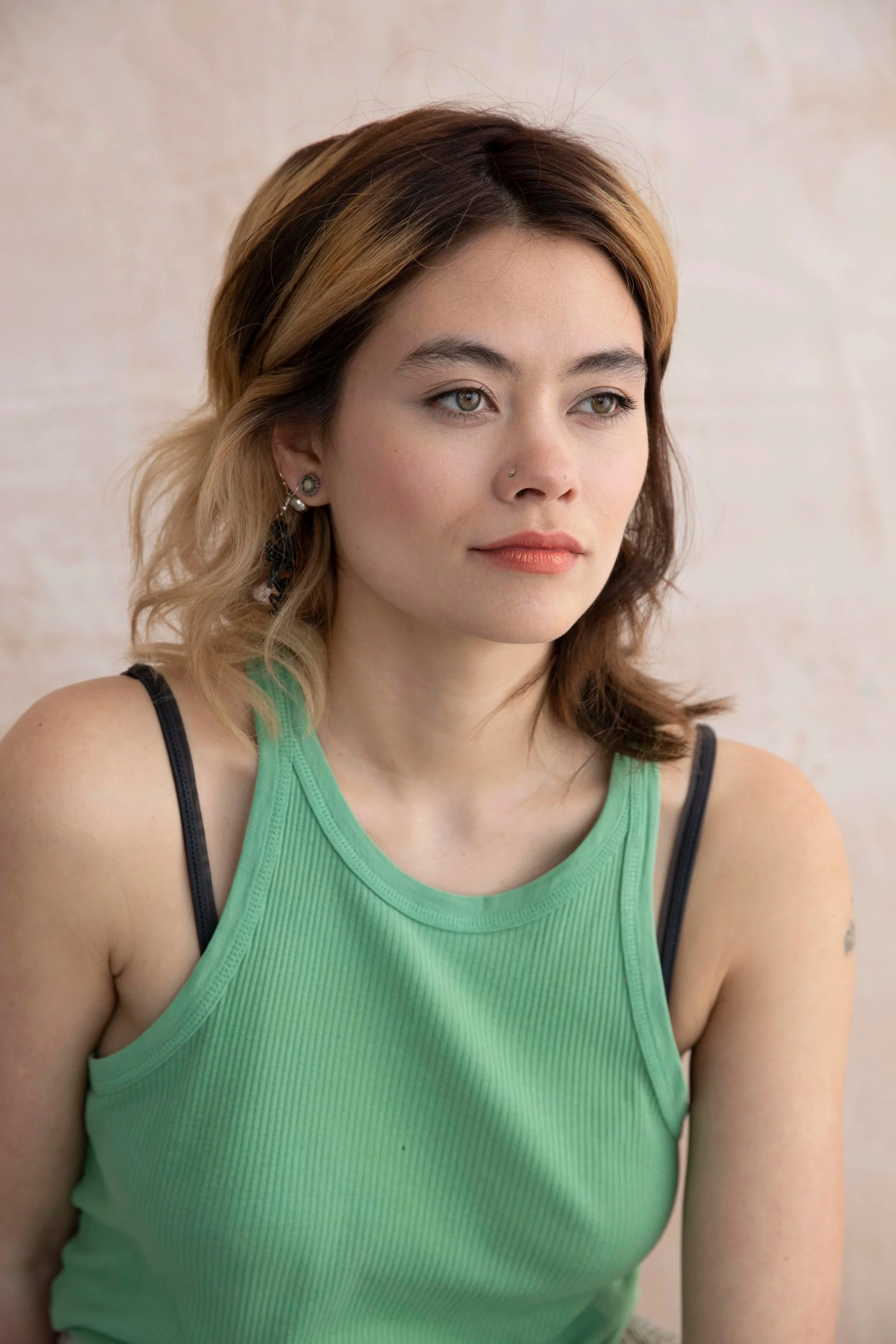 A woman with shoulder-length hair, wearing a green sleeveless top and black straps, looking to the side with a neutral expression against a light-colored backdrop.