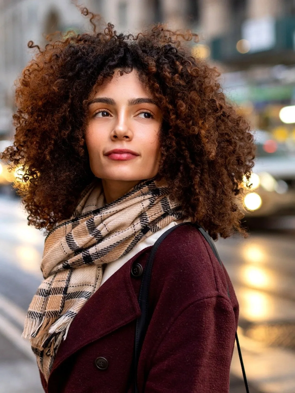 A woman with curly hair walking in a city street during sunset, wearing a plaid scarf and a burgundy coat.