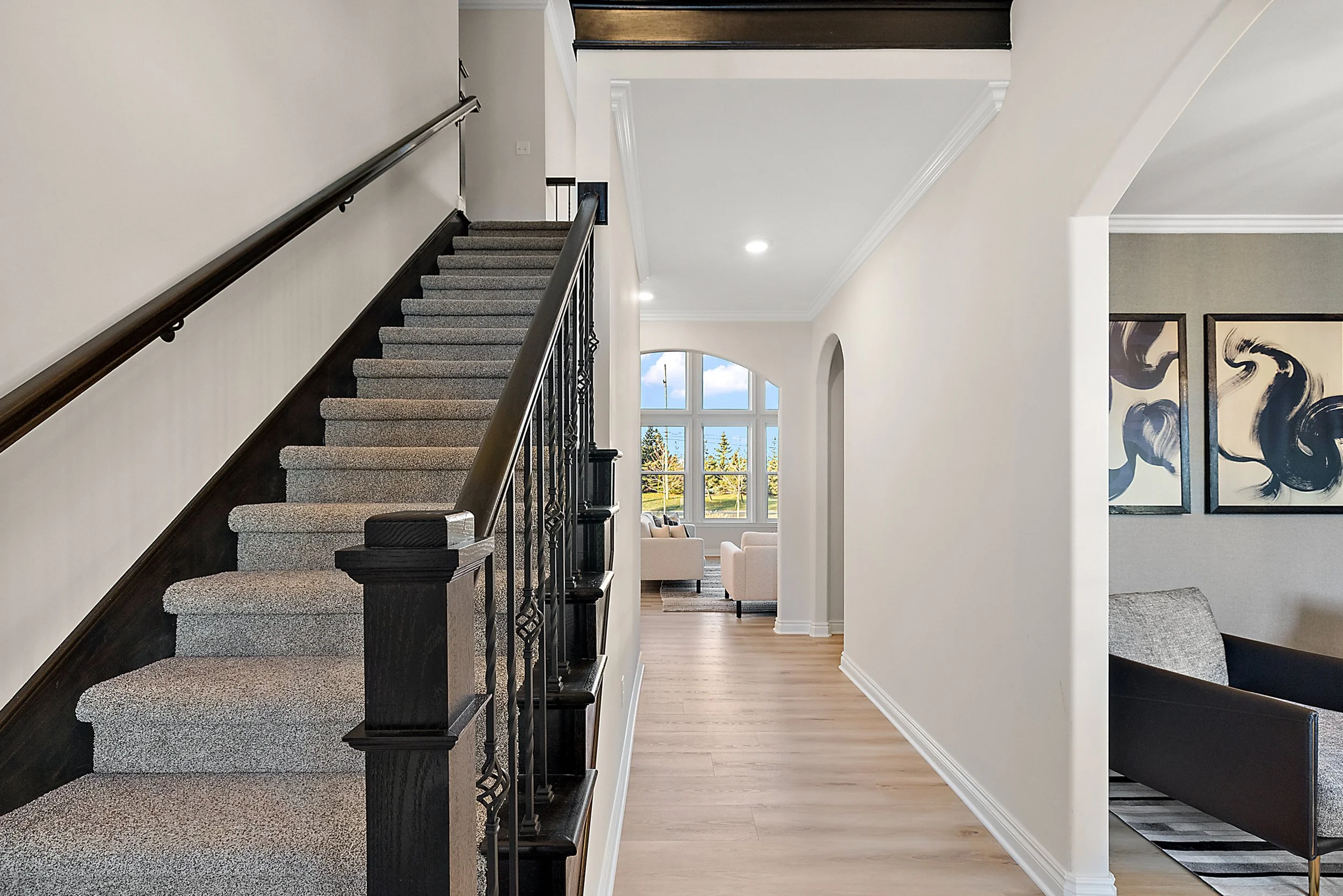 Interior view of a modern home with a staircase on the left, leading upstairs. The hallway has light-colored walls, crown molding, and wood flooring. Visible in the background is a living room with large windows, white sofas, and artwork on the wall.