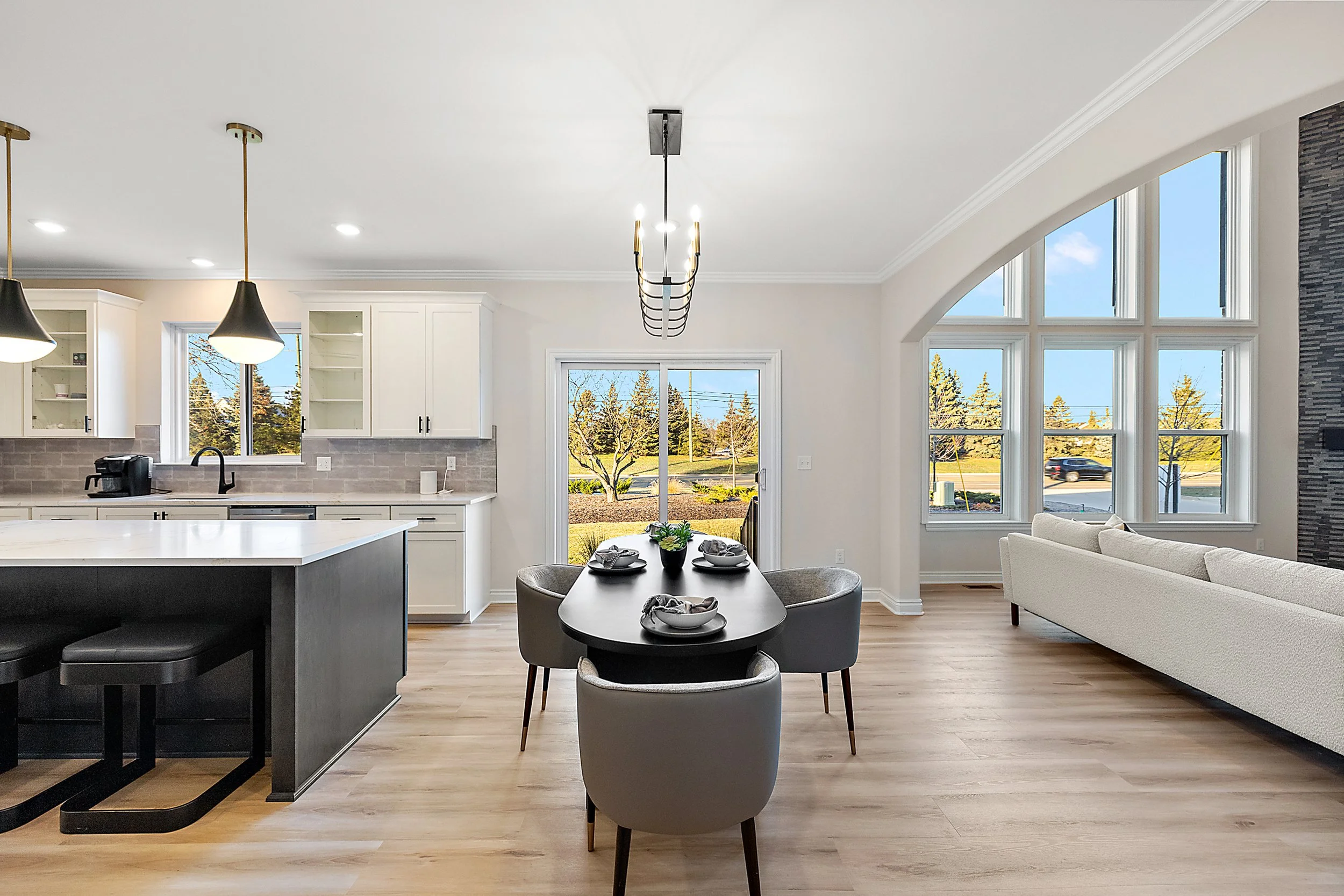 Open-concept living and kitchen area with hardwood floors, large windows, a black dining table with chairs, a white sofa, and a sliding glass door leading outside.