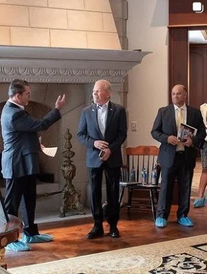 Four men in suits and blue shoe covers standing and talking in a room with a fireplace and a wooden bench.